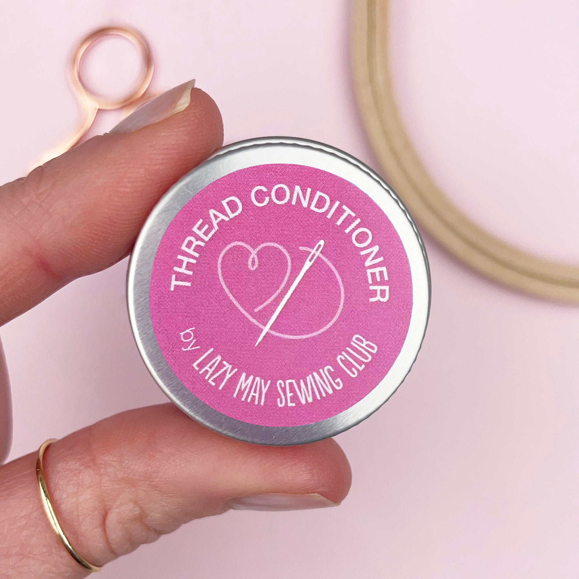 Hand holding a tin labeled 'Thread Conditioner by Lazy May Sewing Club' with sewing tools in the background.