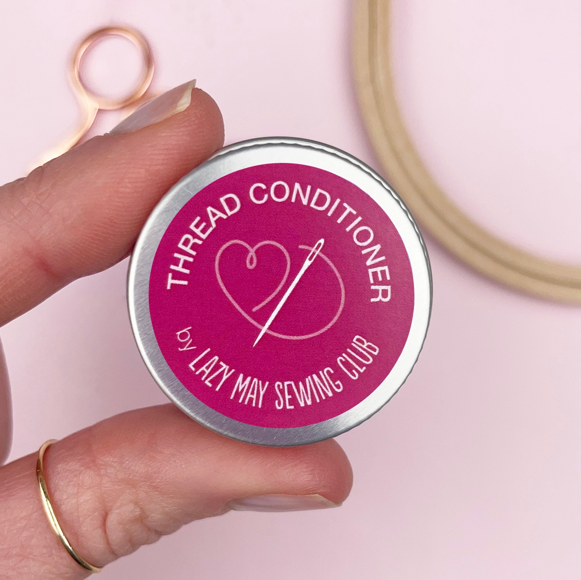 Hand holding a tin labeled 'Thread Conditioner by Lazy May Sewing Club' with sewing tools in the background.