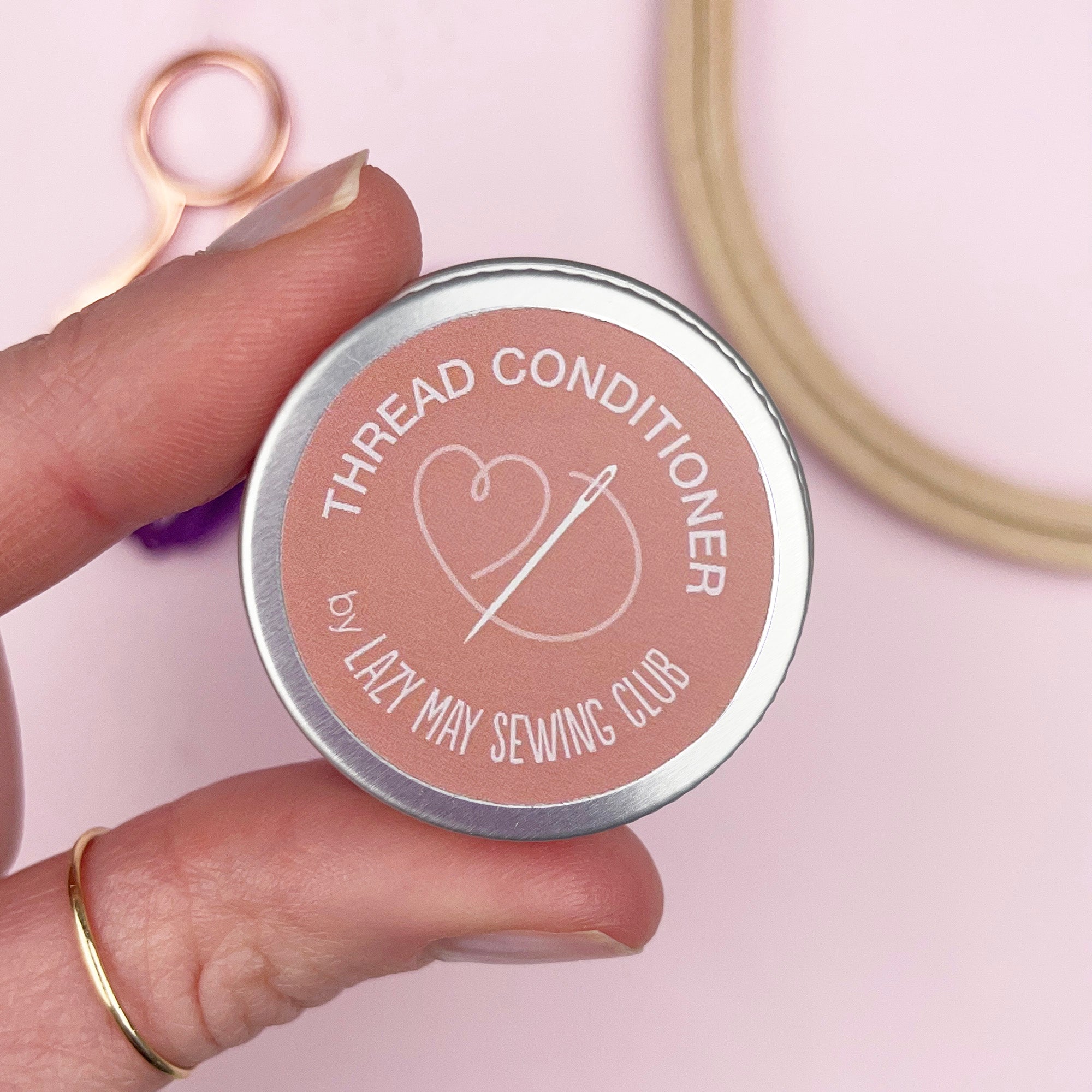 Hand holding a 'Thread Conditioner' container by Lazy May Sewing Club with sewing tools in the background.