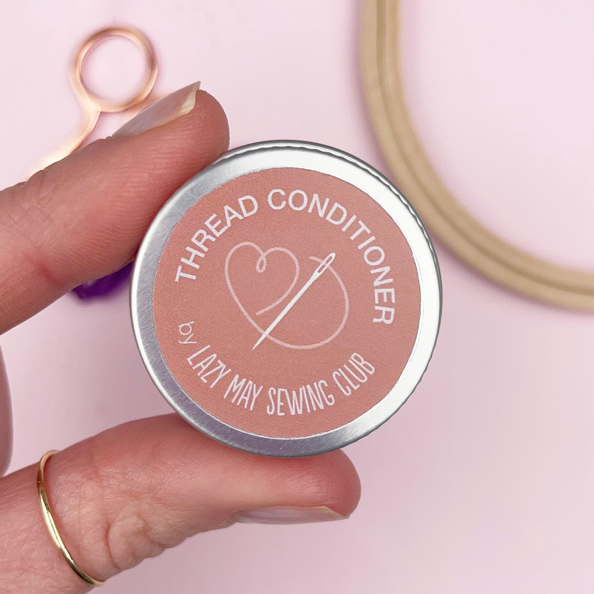 Hand holding a 'Thread Conditioner' container by Lazy May Sewing Club with sewing tools in the background.