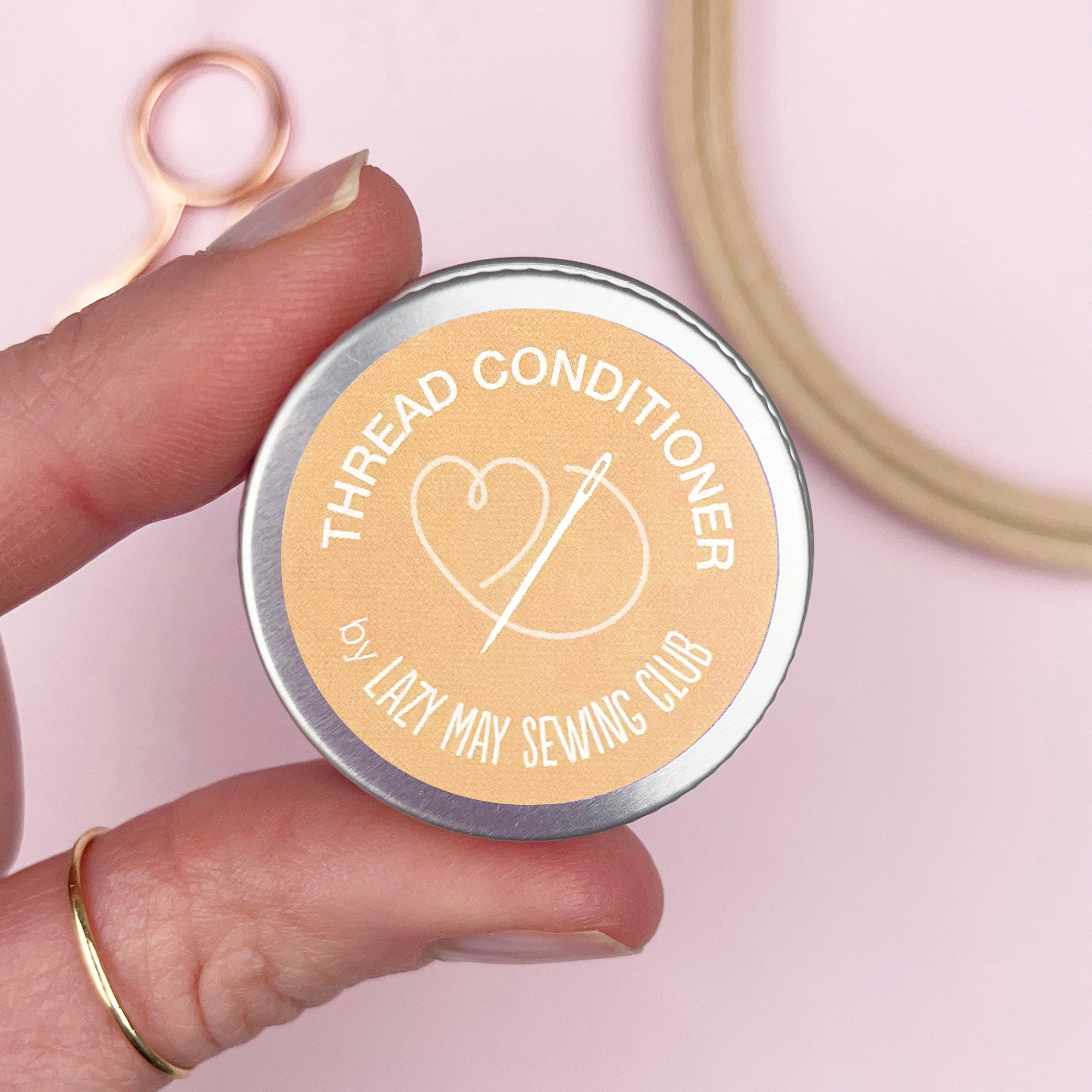 Hand holding a tin labeled 'Thread Conditioner' by Lazy May Sewing Club on a pink background.