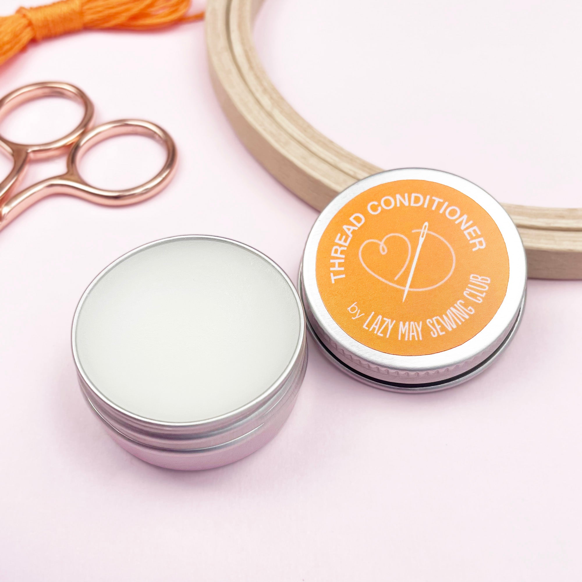 Thread conditioner container with a lid, scissors, and spool of thread on a light pink background