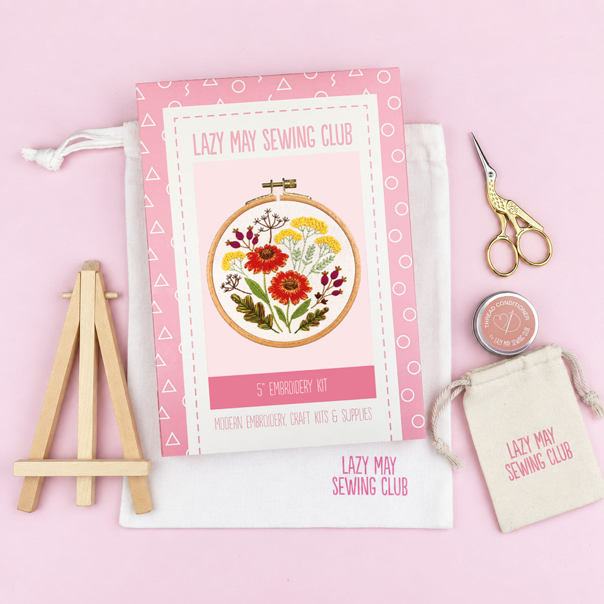 A flat-lay of a 'Lazy May Sewing Club' 5-star embroidery kit on a light pink background. The kit includes a pink and white patterned booklet with a floral embroidery hoop illustration, a white drawstring bag, a small wooden easel, gold scissors, and a small round tin of thread.