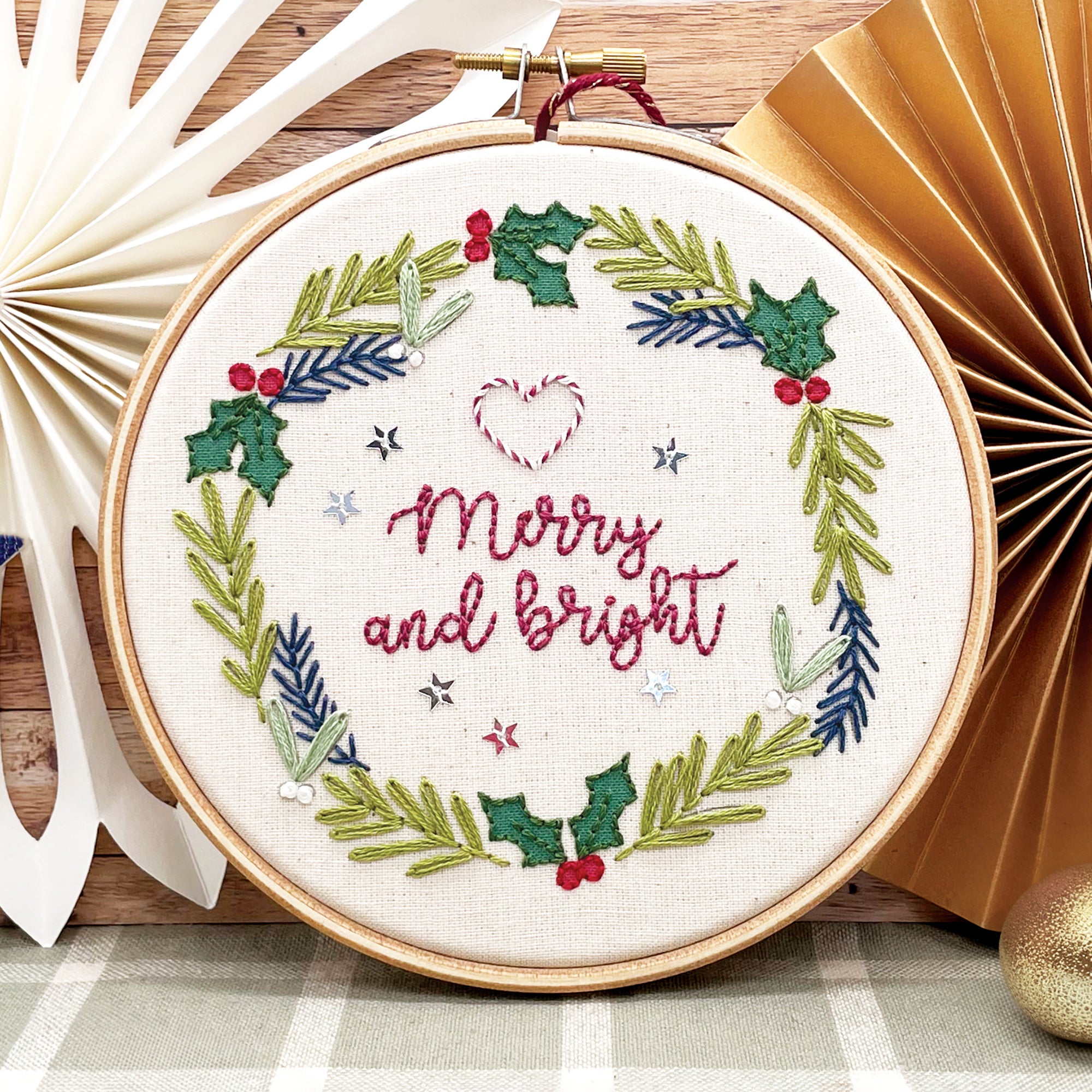 A festive Christmas wreath embroidery kit displayed in a hoop, featuring red text saying 'Merry and bright', surrounded by green holly and berries.