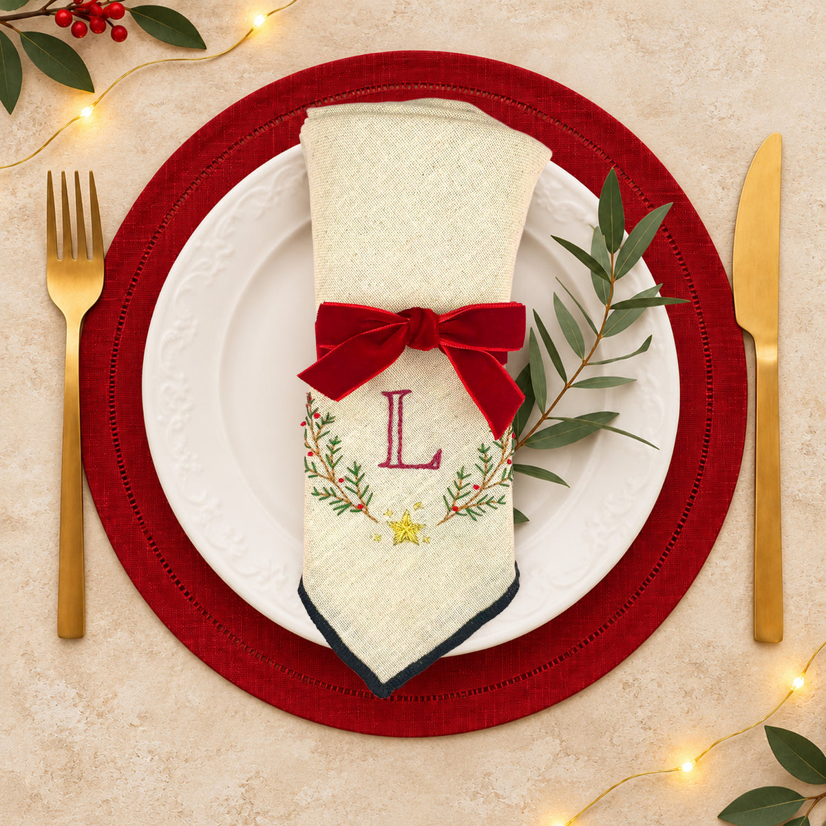 A festive table setting featuring a cream-colored napkin with a red velvet bow, embroidered monogram 'L', and holiday wreath details, styled on a white plate with a red charger placemat, gold cutlery, and green leaves.