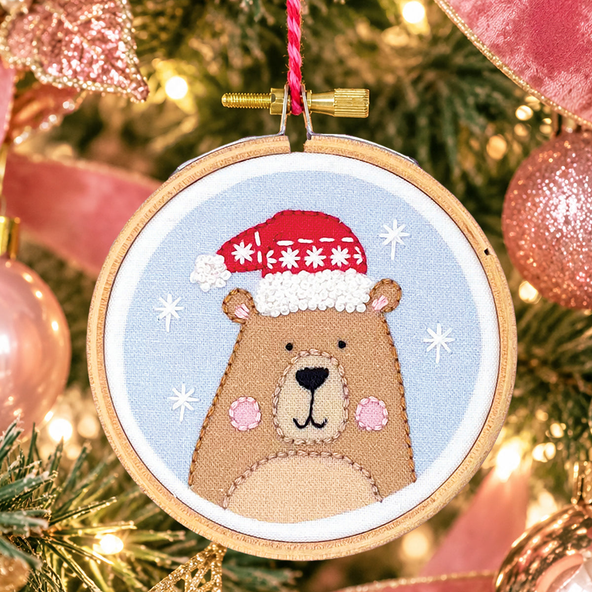 A circular embroidery hoop ornament featuring a stitched brown bear wearing a red Santa hat on a light blue background, hanging in front of a softly blurred Christmas tree with pink ornaments and warm lights.
