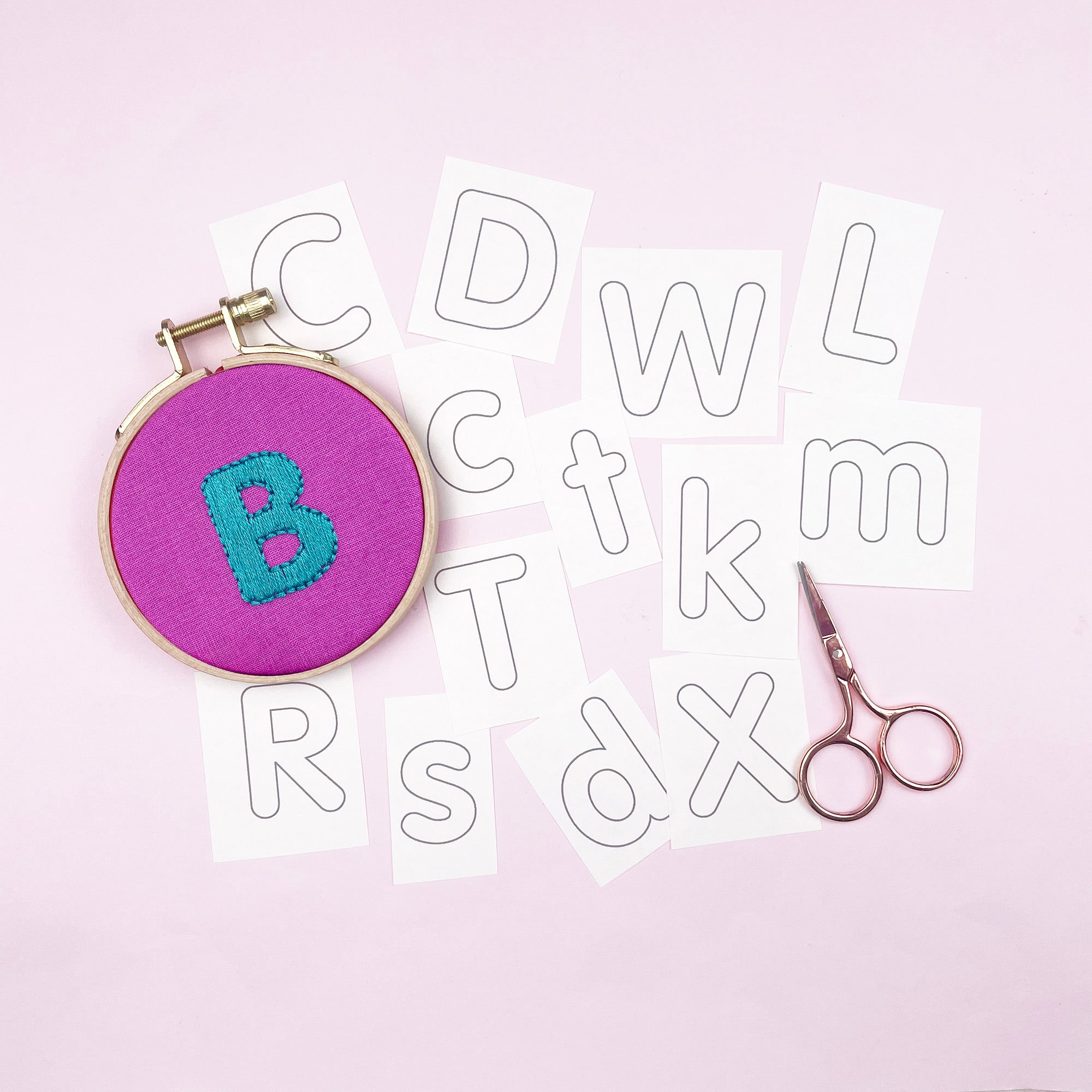 Embroidery hoop with letter 'B' on a pink background with cut-out iron on embroidery patterns of letters and scissors.
