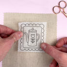Hands holding a fabric swatch with a printed design of a teapot and tulip, next to a pair of scissors on a light pink background.