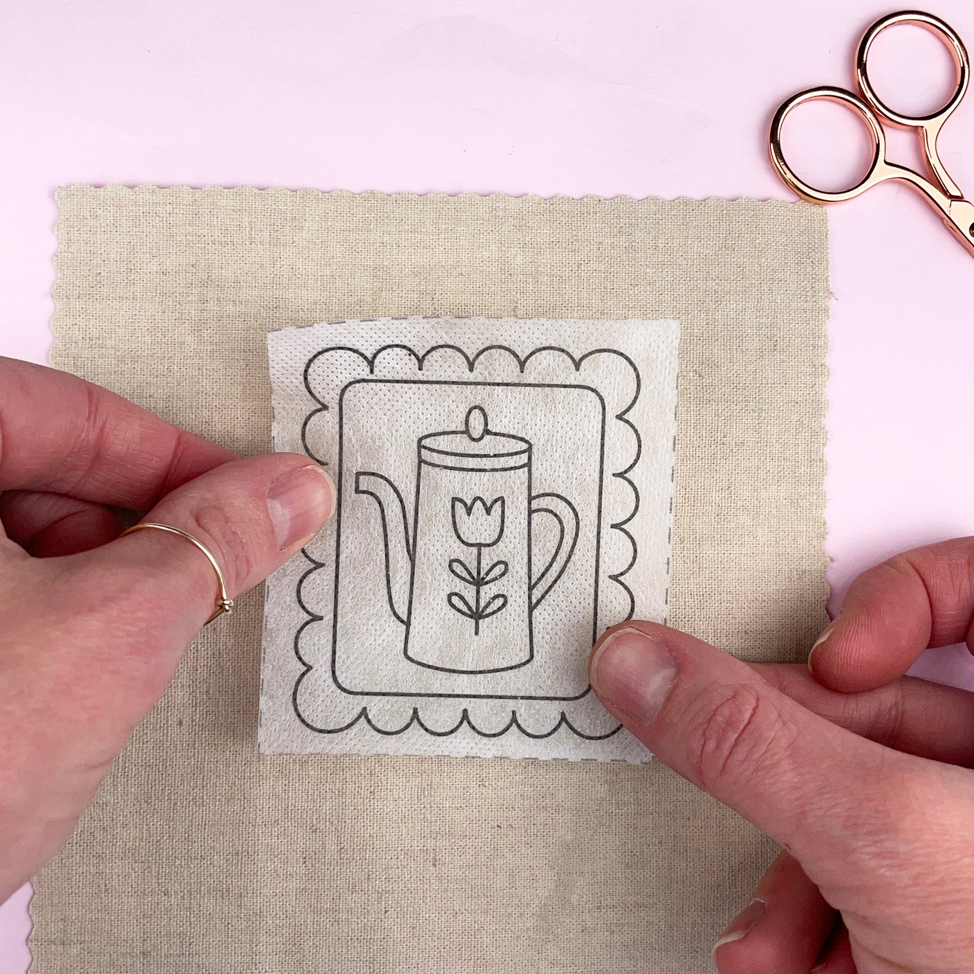 Hands holding a fabric swatch with a printed design of a teapot and tulip, next to a pair of scissors on a light pink background.