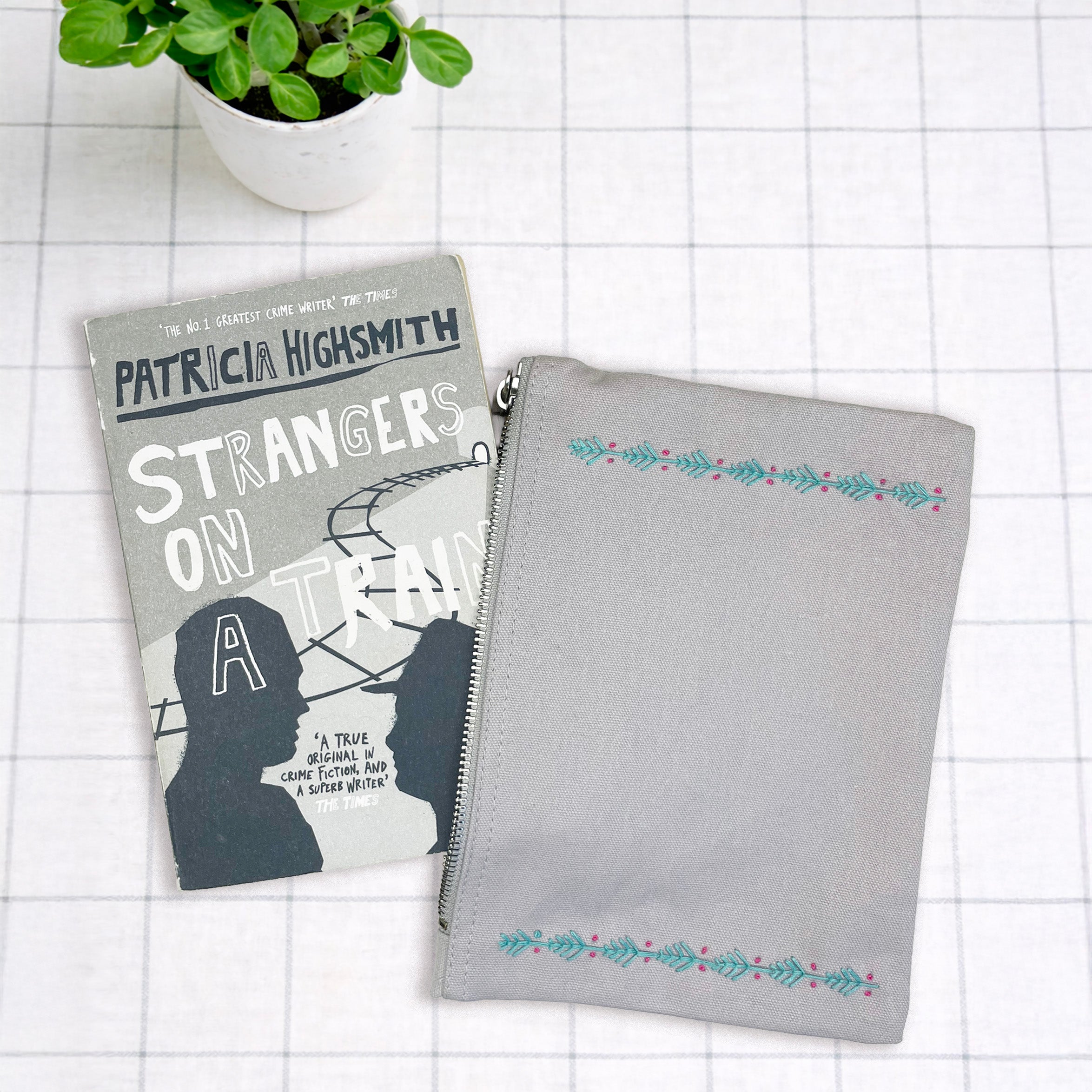 Book titled 'Strangers on a Train' by Patricia Highsmith next to a gray pouch with floral embroidery on a white grid background.