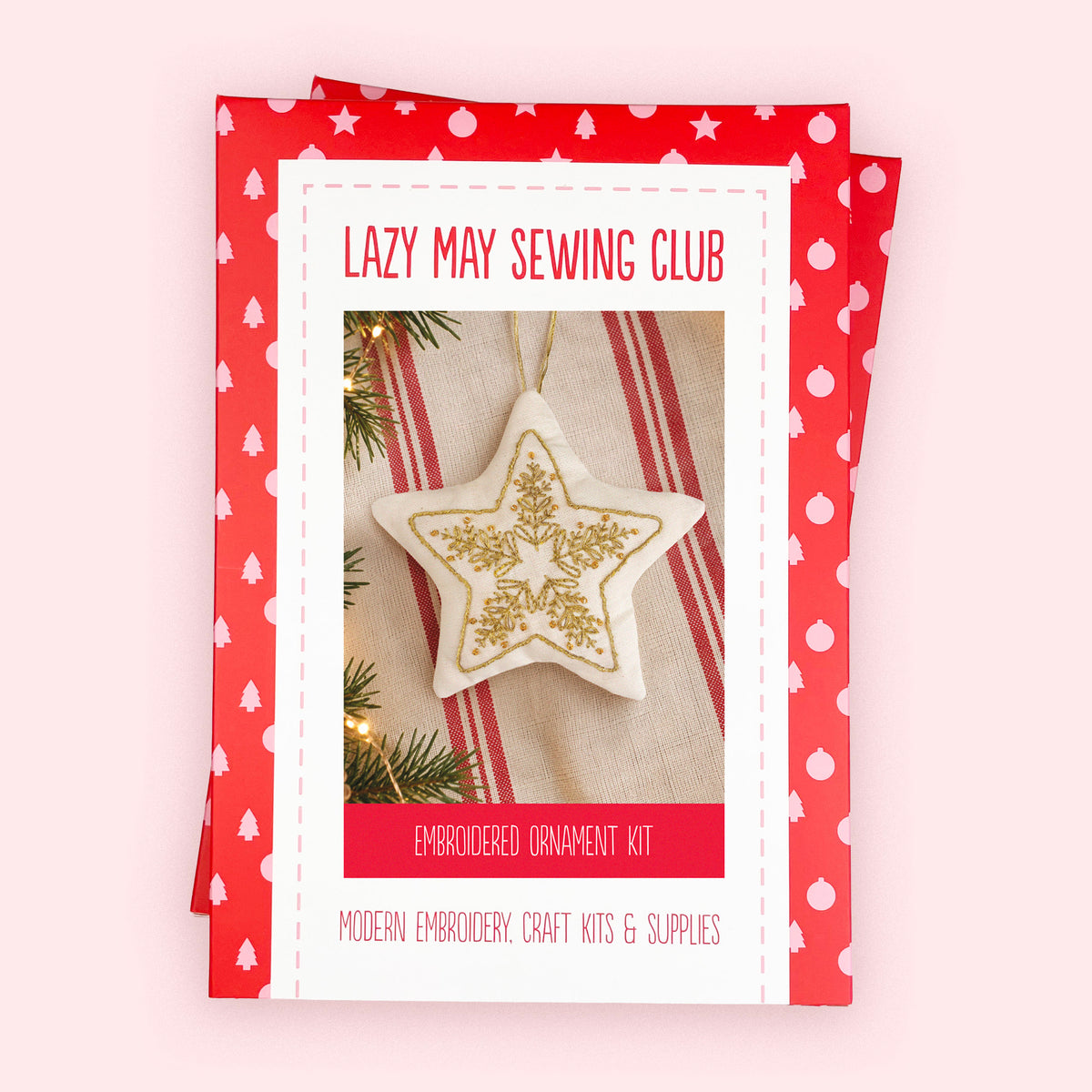A boxed embroidery kit package titled 'Lazy May Sewing Club' with a red-and-white polka-dot border and a central photo of a star-shaped embroidered ornament. The package is photographed on a pale pink background.