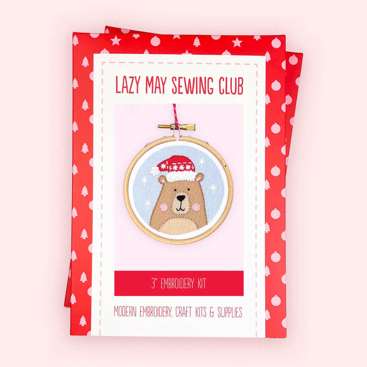 A 3" embroidery kit package from Lazy May Sewing Club, featuring a cartoon bear wearing a Santa hat inside an embroidery hoop illustration on a white card with red borders, set against a light pink background.
