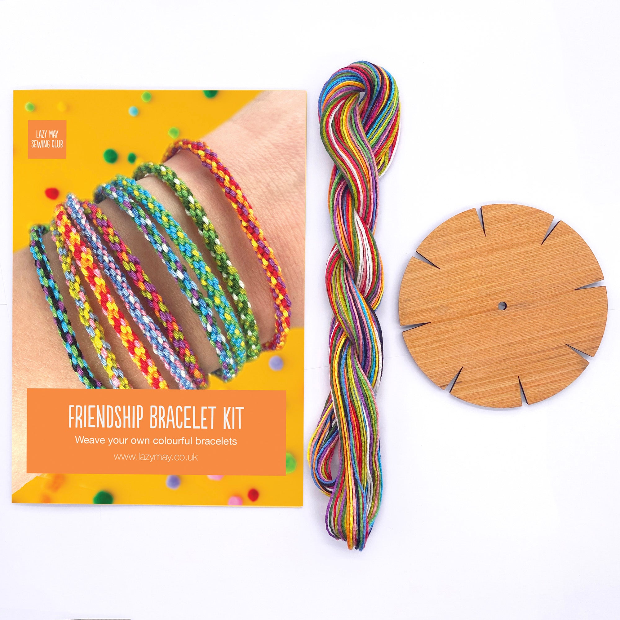 The full rainbow bracelet making kit laid out – includes a round wooden kumihimo loom, a bundle of rainbow threads, and a printed instruction card on a bright background.