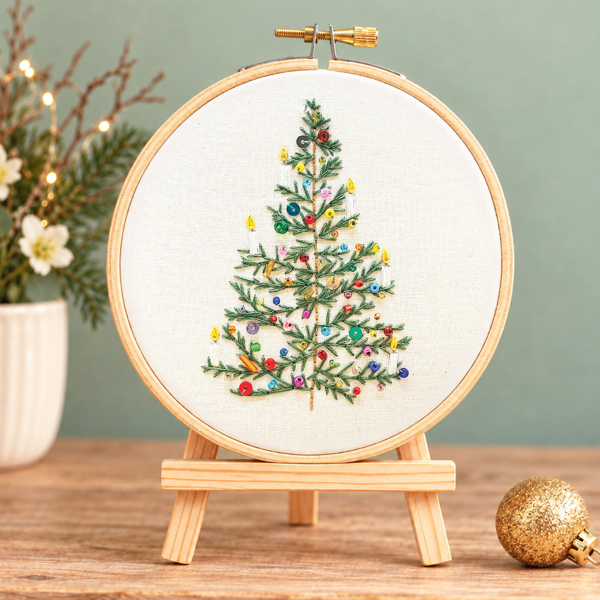 A finished embroidery piece showing a decorated Christmas tree inside a wooden hoop on a small wooden stand, set on a tabletop with a blurred holiday scene in the background.