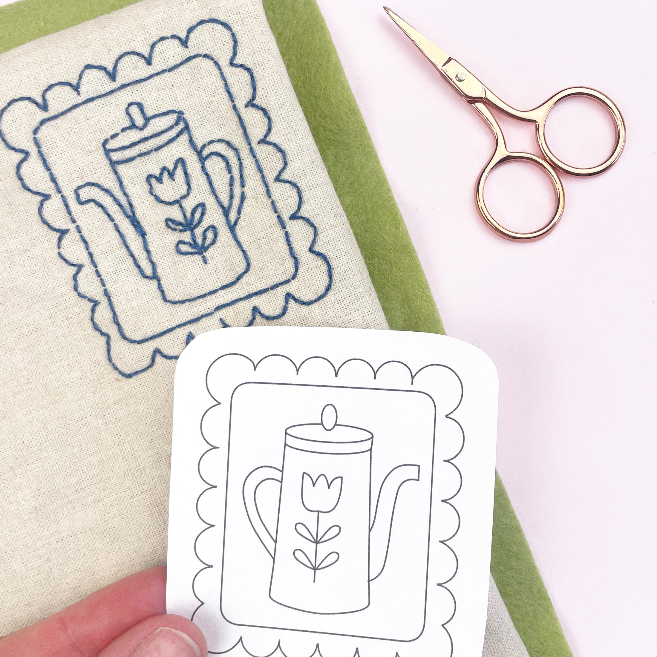 Embroidery pattern card with a teapot design held by a hand, next to a piece of fabric with embroidery and a pair of scissors.