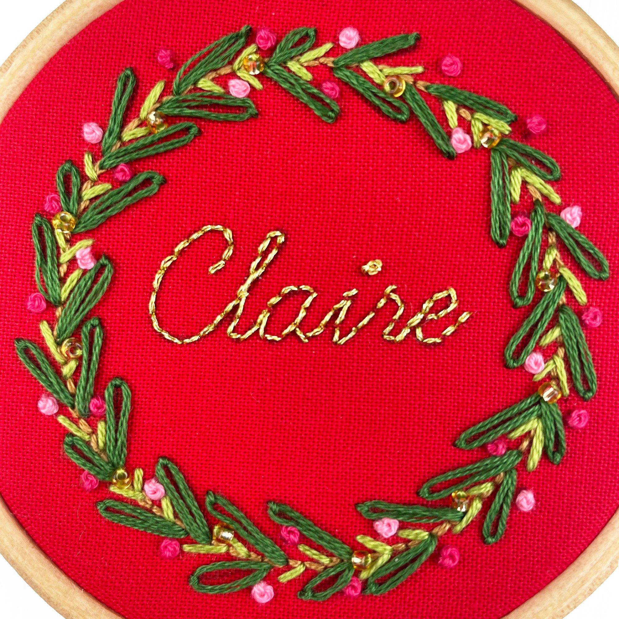 Embroidery hoop with red fabric, green and gold thread, and pink beads spelling 'Claire'.