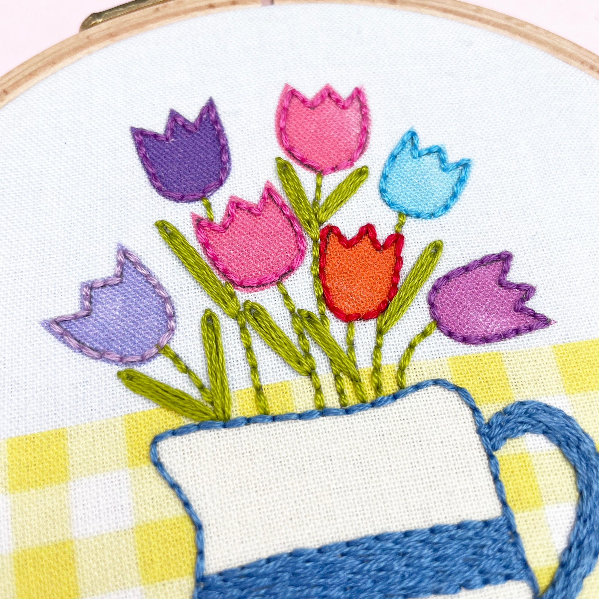 Embroidery hoop with colorful tulips in a blue and white pitcher on a yellow checkered background.