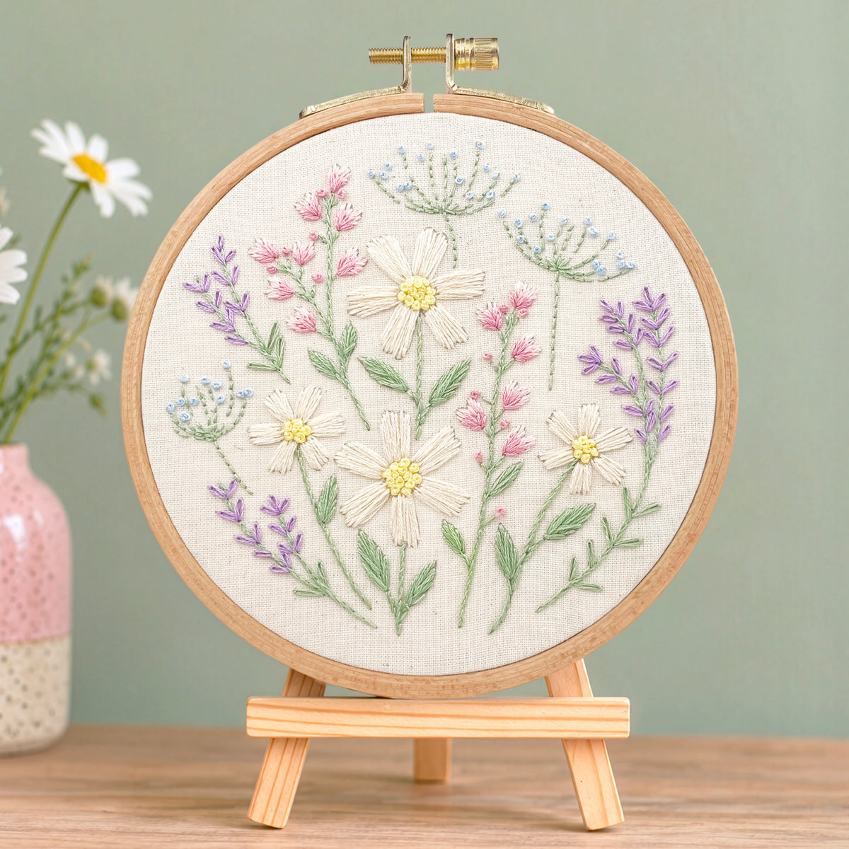 A round embroidery hoop holding cream fabric with embroidered wildflowers in white, pink, purple, and blue, displayed on a small wooden stand against a soft green wall with a vase of daisies in the background.