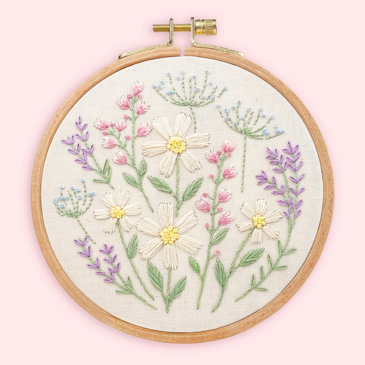 A round wooden embroidery hoop holding a floral embroidered textile design on a pale pink background. The embroidery features cream daisies with yellow centers, pink blossoms, green stems and leaves, and small blue flower clusters.