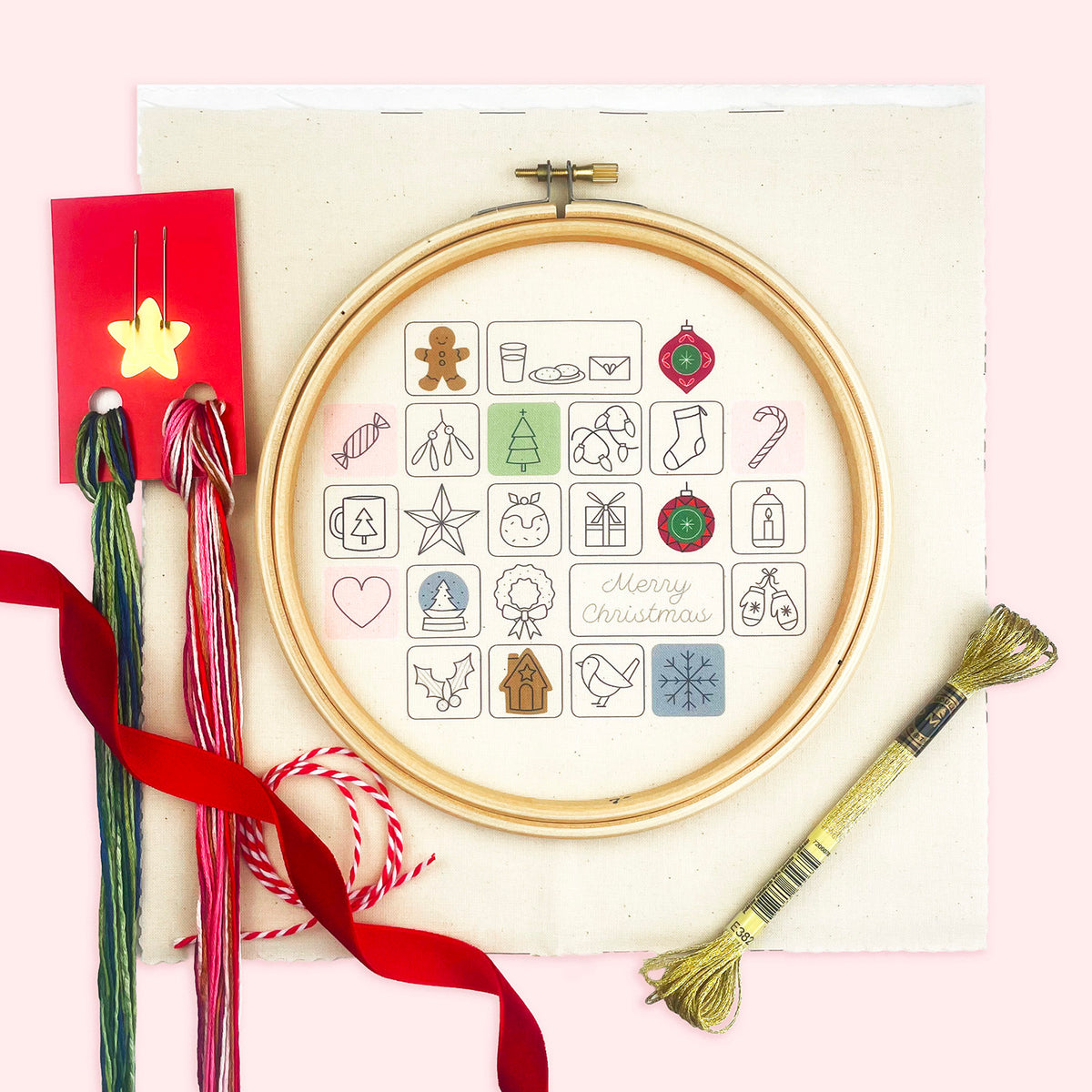 A circular embroidery hoop advent calendar kit with Christmas-themed design on cream fabric, surrounded by crafting props including red ribbon, embroidery floss, a red card with a star, and a gold thread bundle, all arranged on a light pink background.
