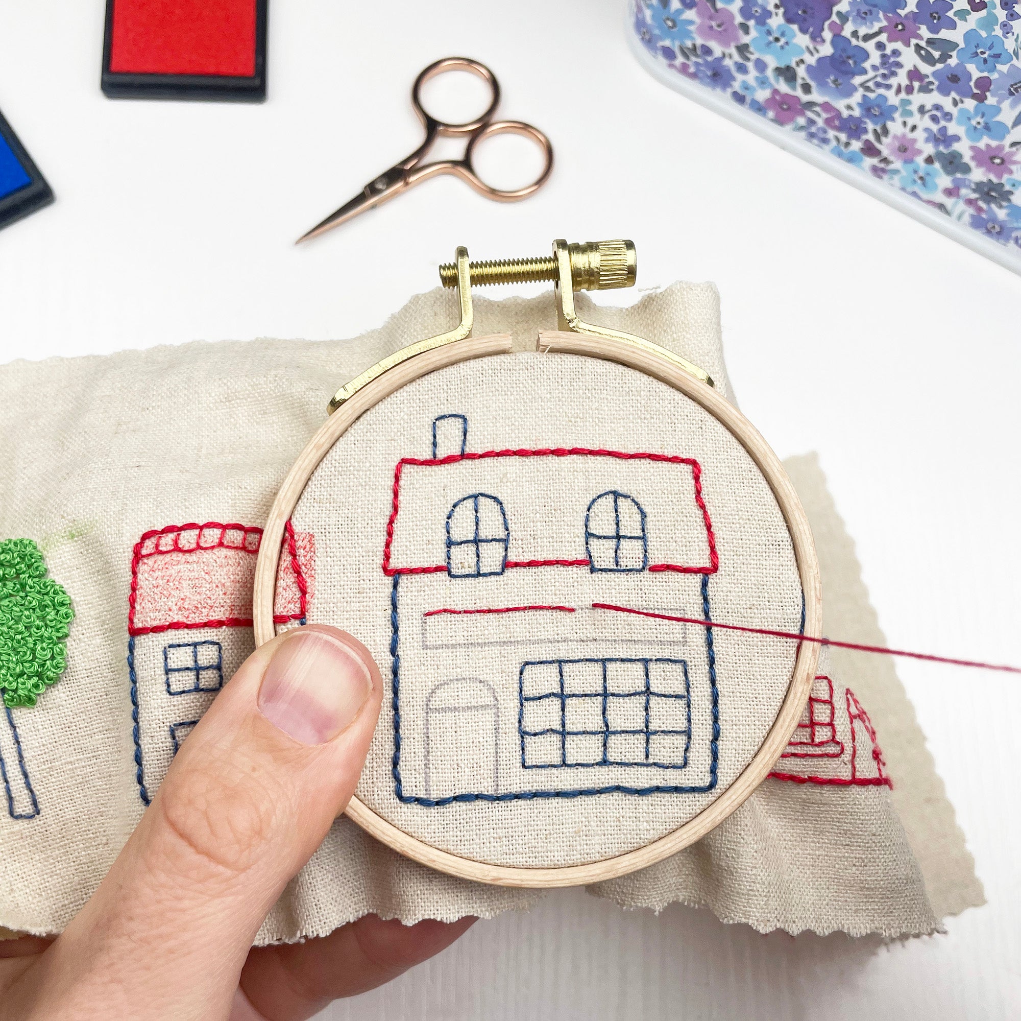 Embroidery hoop with a house design on fabric, surrounded by sewing tools on a white surface. iron on embroidery patterns
