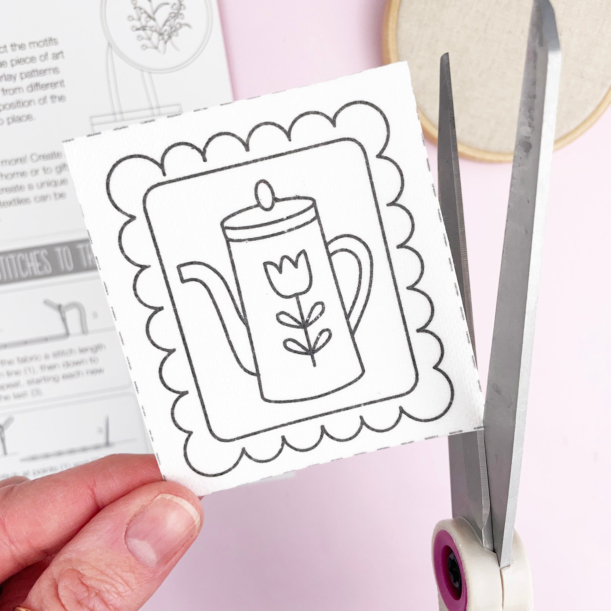 Hand holding a card with a black and white stick and stitch embriodery pattern of a mug and flower, scissors nearby on a light purple background.