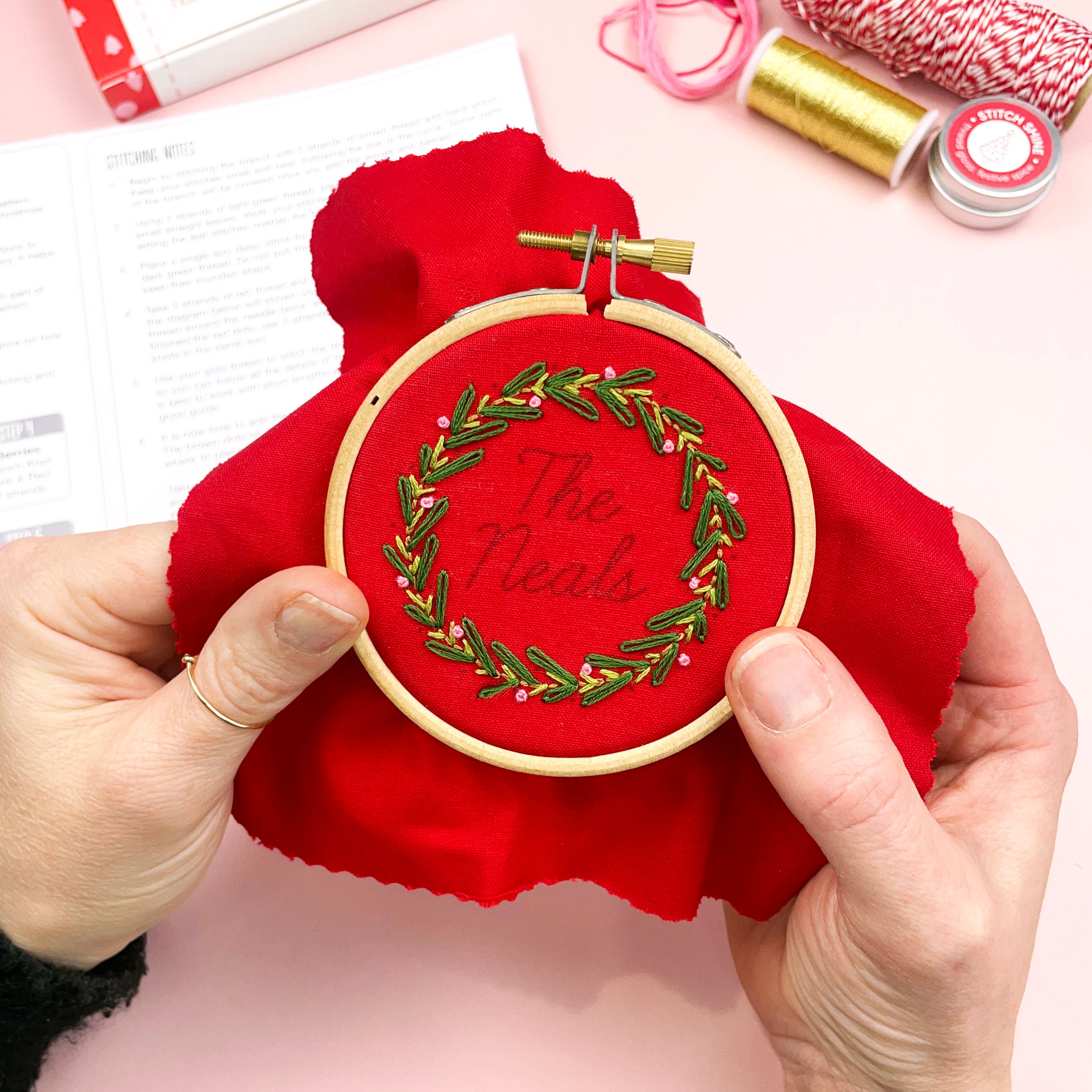 Embroidery hoop with red fabric and embroidered design held by hands on a pink surface.