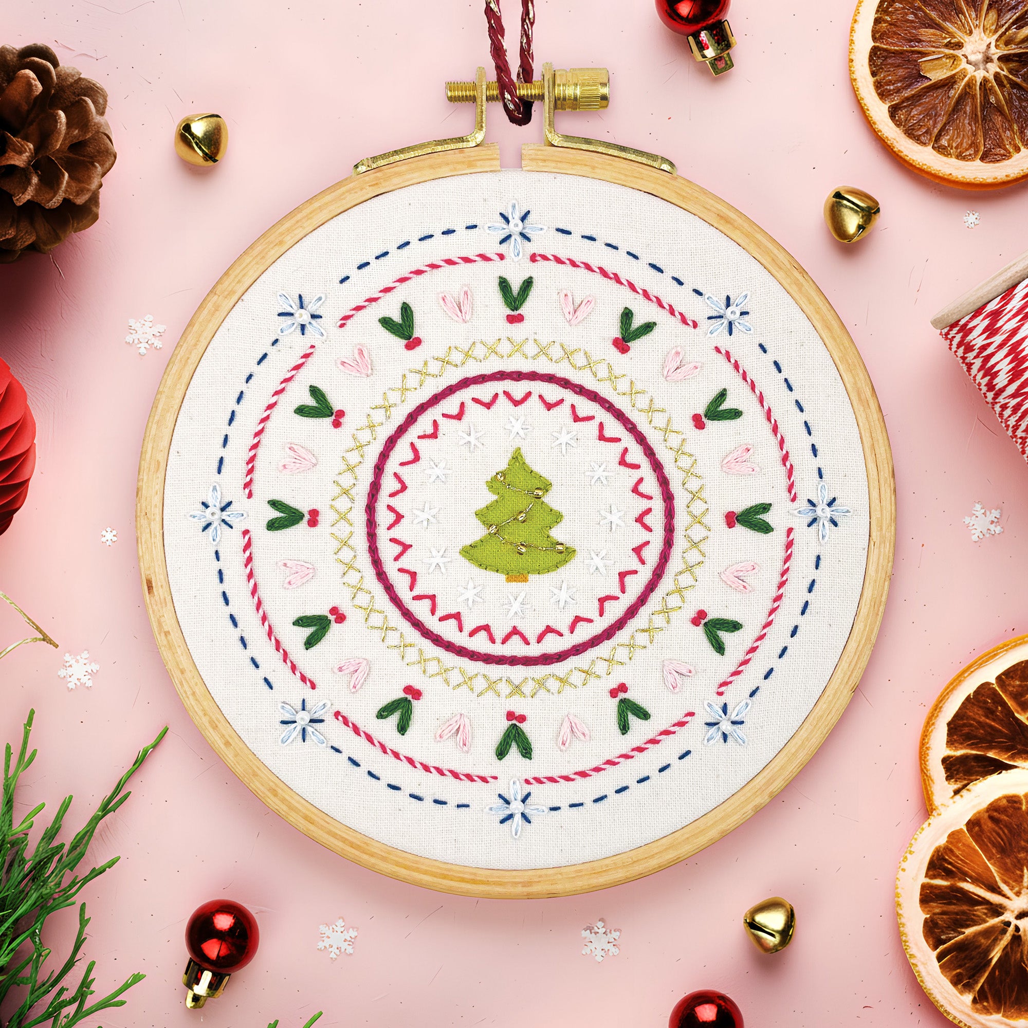 Embroidery hoop with Christmas-themed design on a pink background with festive decorations.