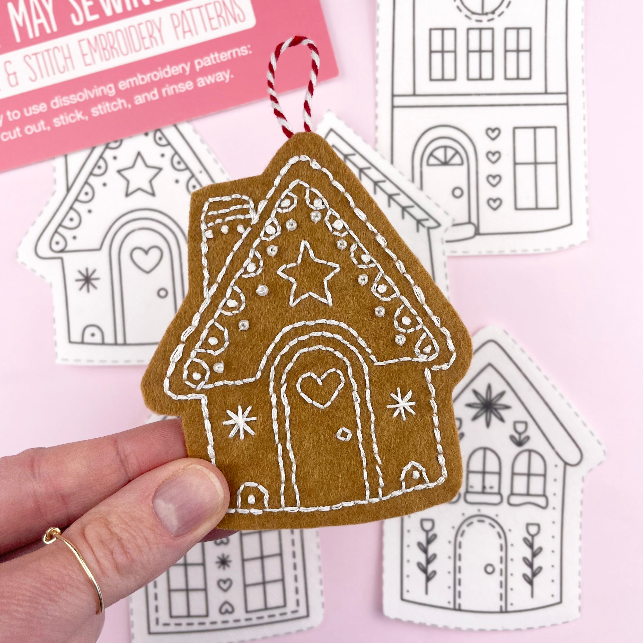 Hand holding a gingerbread house-shaped embroidered ornament with sewing patterns in the background.
