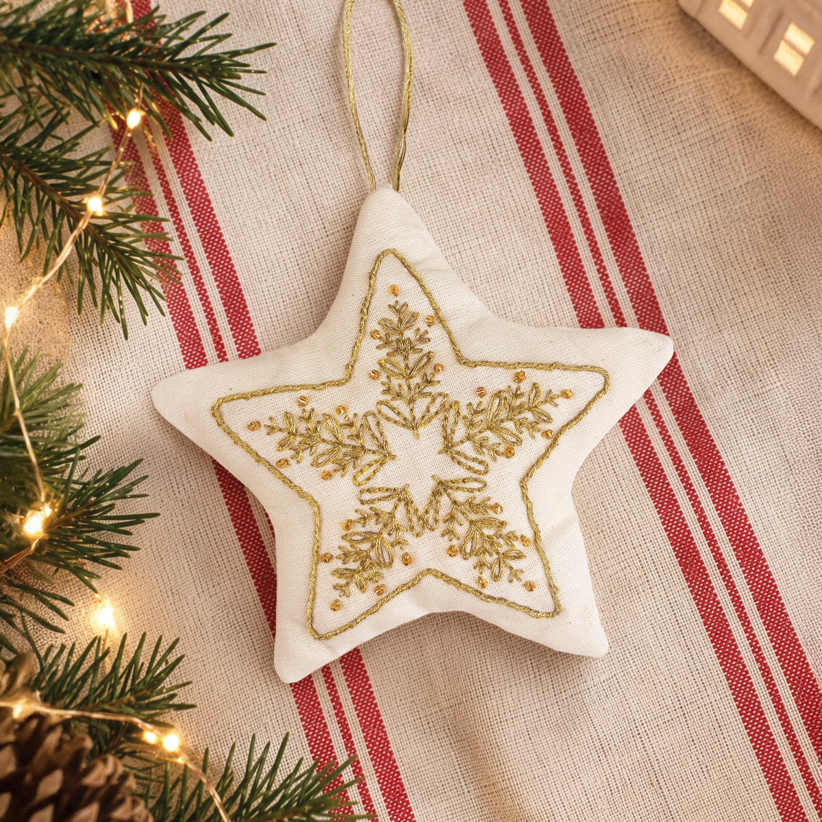 A star-shaped white fabric ornament with a gold embroidered snowflake design, hanging on a red and beige striped textile with pine branches and warm string lights around it.
