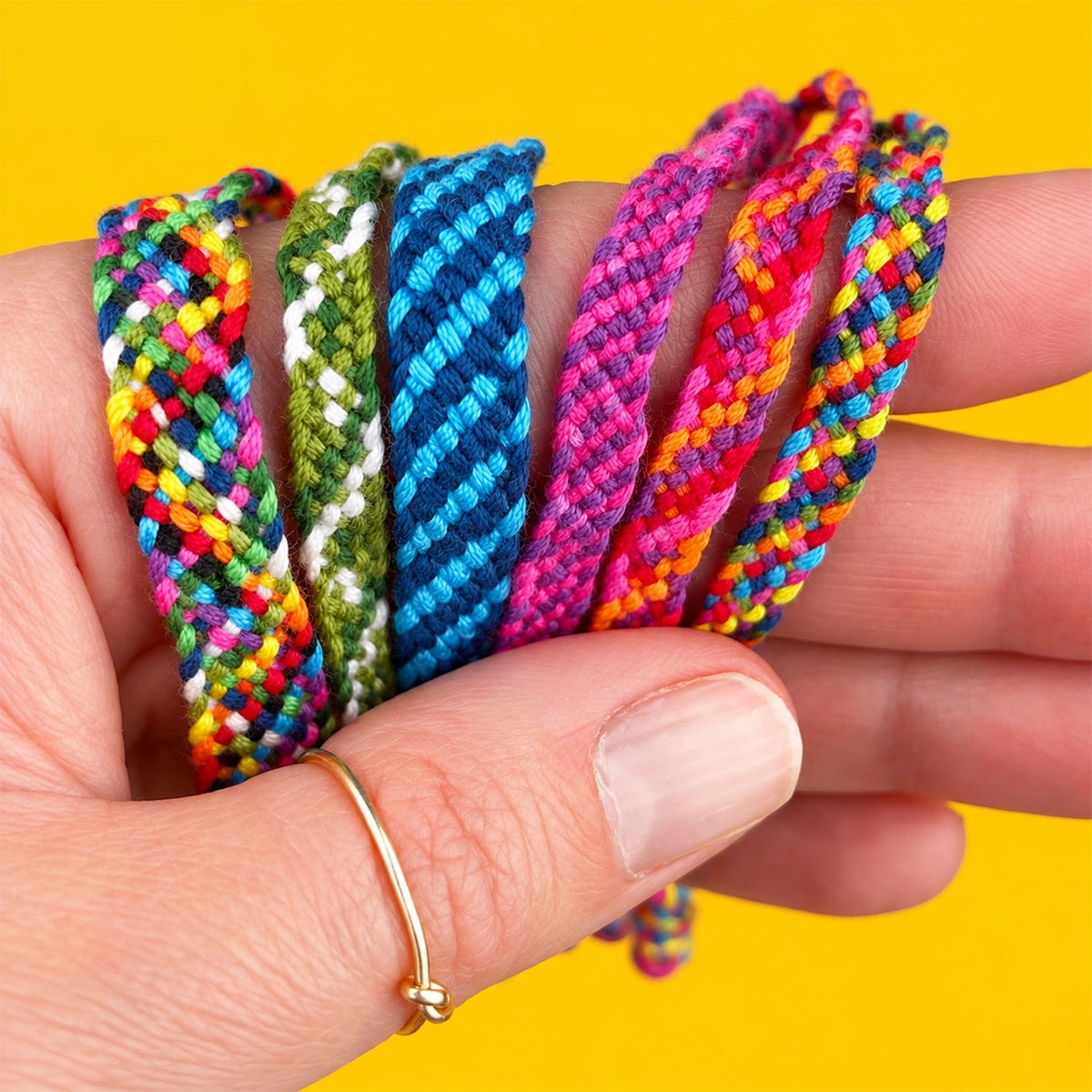 A close-up of a hand holding five braided friendship bracelets in various vibrant colors, including yellow, green, blue, pink, and multicolor, set against a solid bright yellow background.