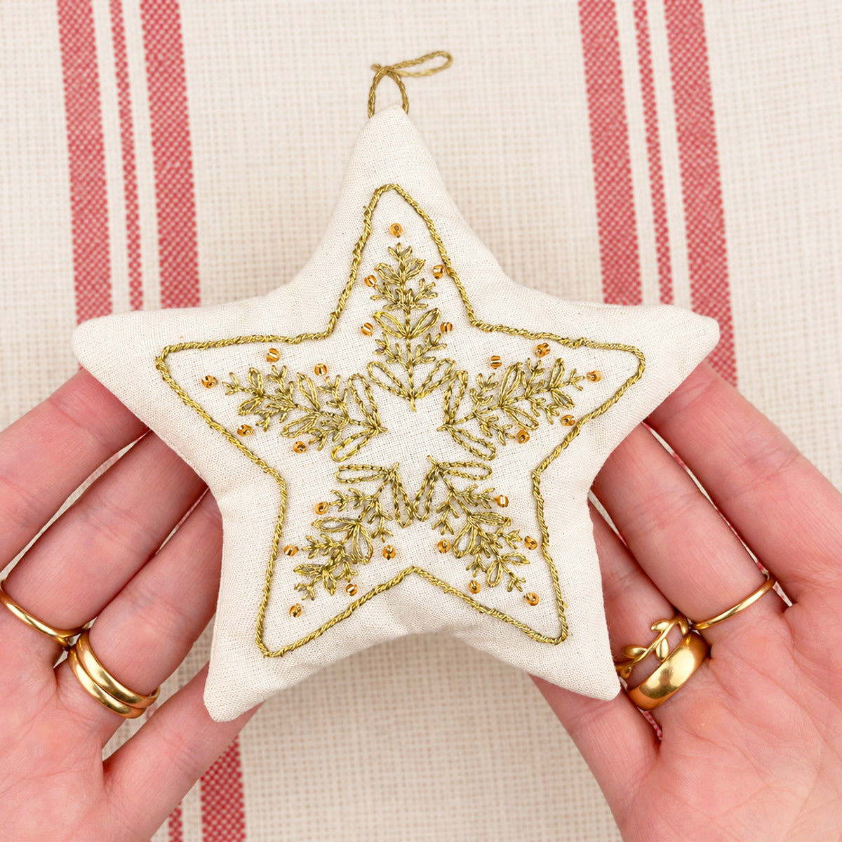 A hand holds a star-shaped cream fabric ornament with gold embroidered snowflake and pine branch patterns, against a red and white striped fabric background.