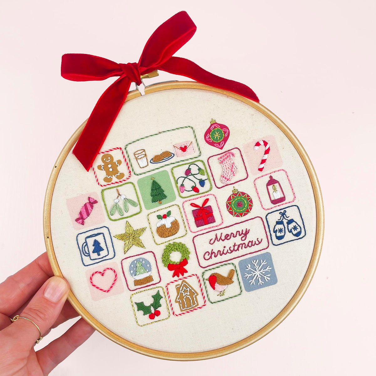 A hand holds a wooden embroidery hoop advent calendar containing a completed Christmas-themed embroidery design with embroidered icons, a red ribbon bow, and a 'Merry Christmas' message on a light pink background.