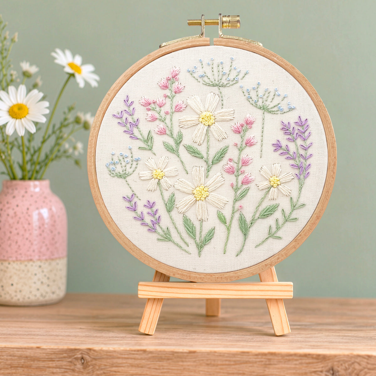 A round embroidery hoop holding a floral embroidered design on cream fabric, displayed on a small wooden stand. The embroidery features white daisies, pink and purple flowers, and green stems on a light wood background.