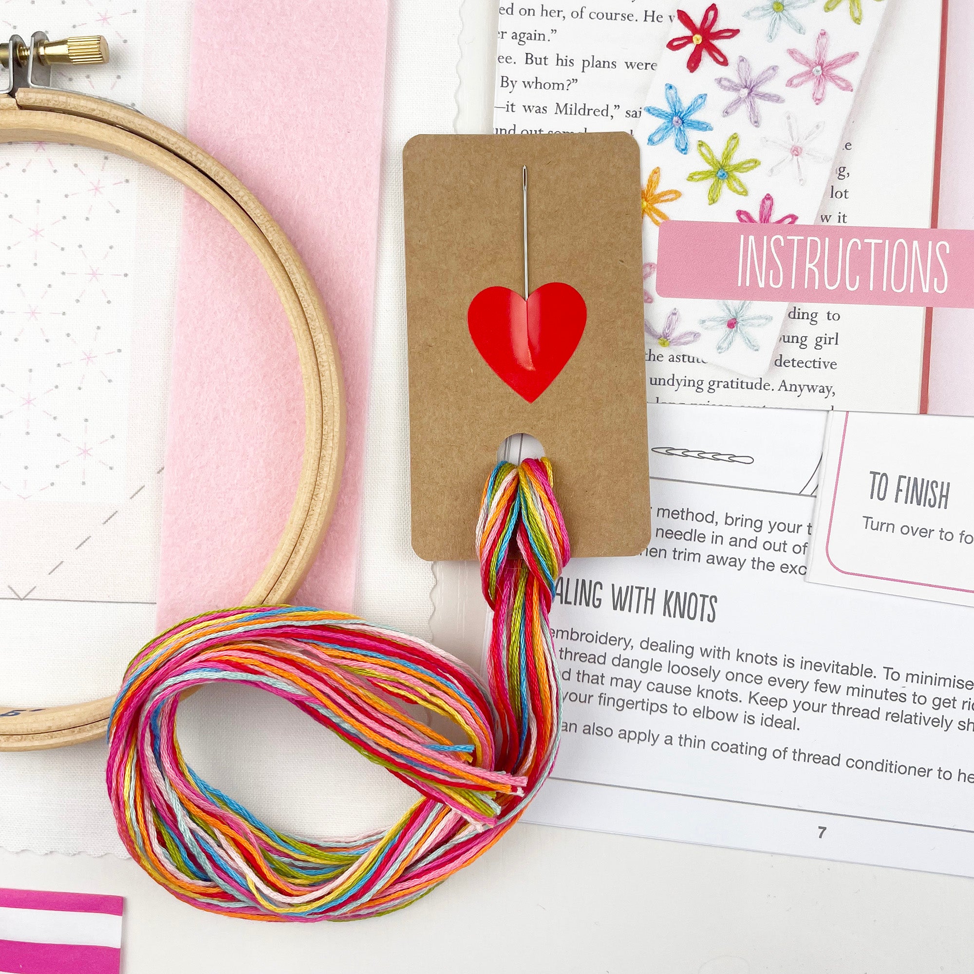 Colorful thread ball with a heart tag on a sewing book and hoop, contents of Hand embroidered bookmark making kit