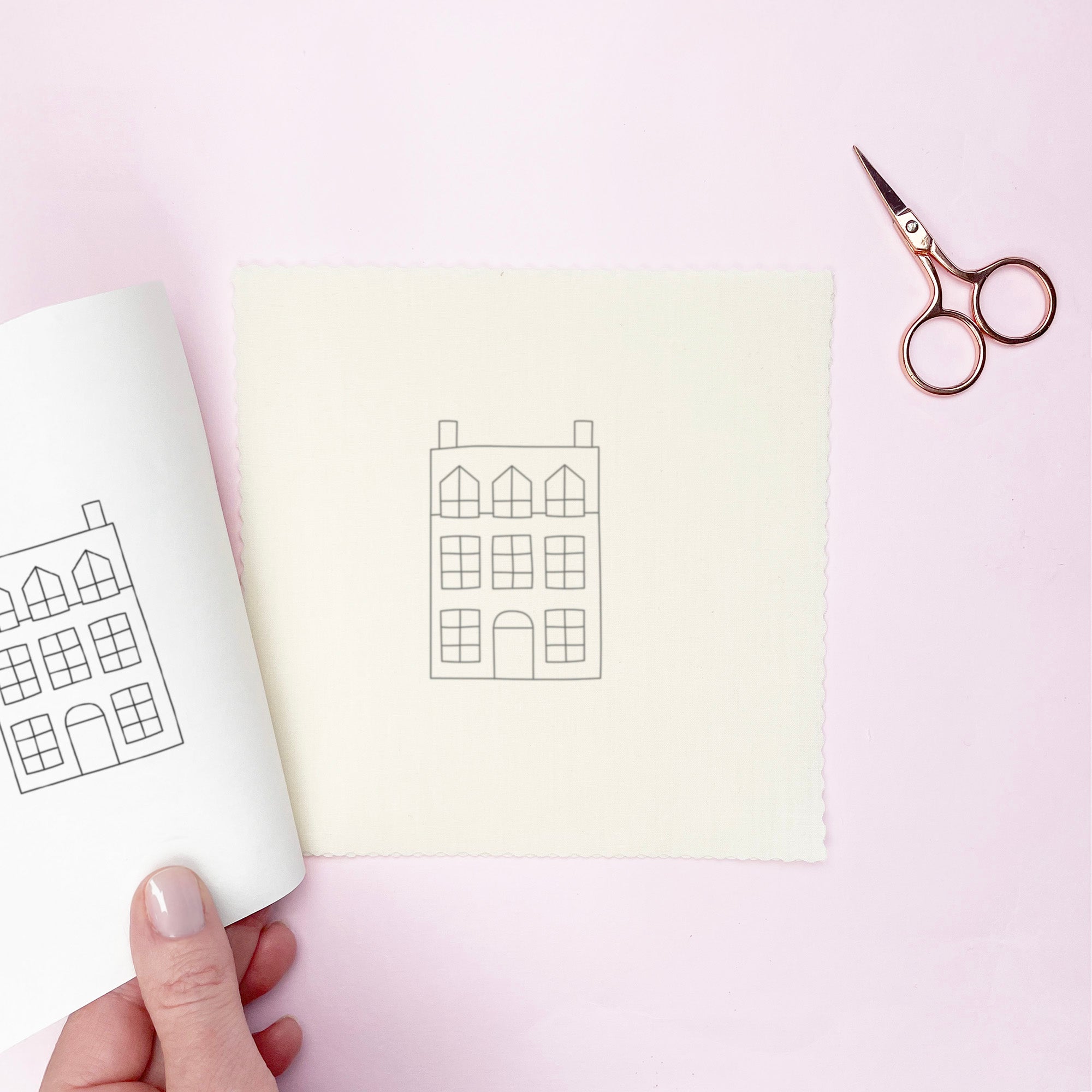 House embroidery pattern cutout and transferred on to fabric on a pink background with scissors