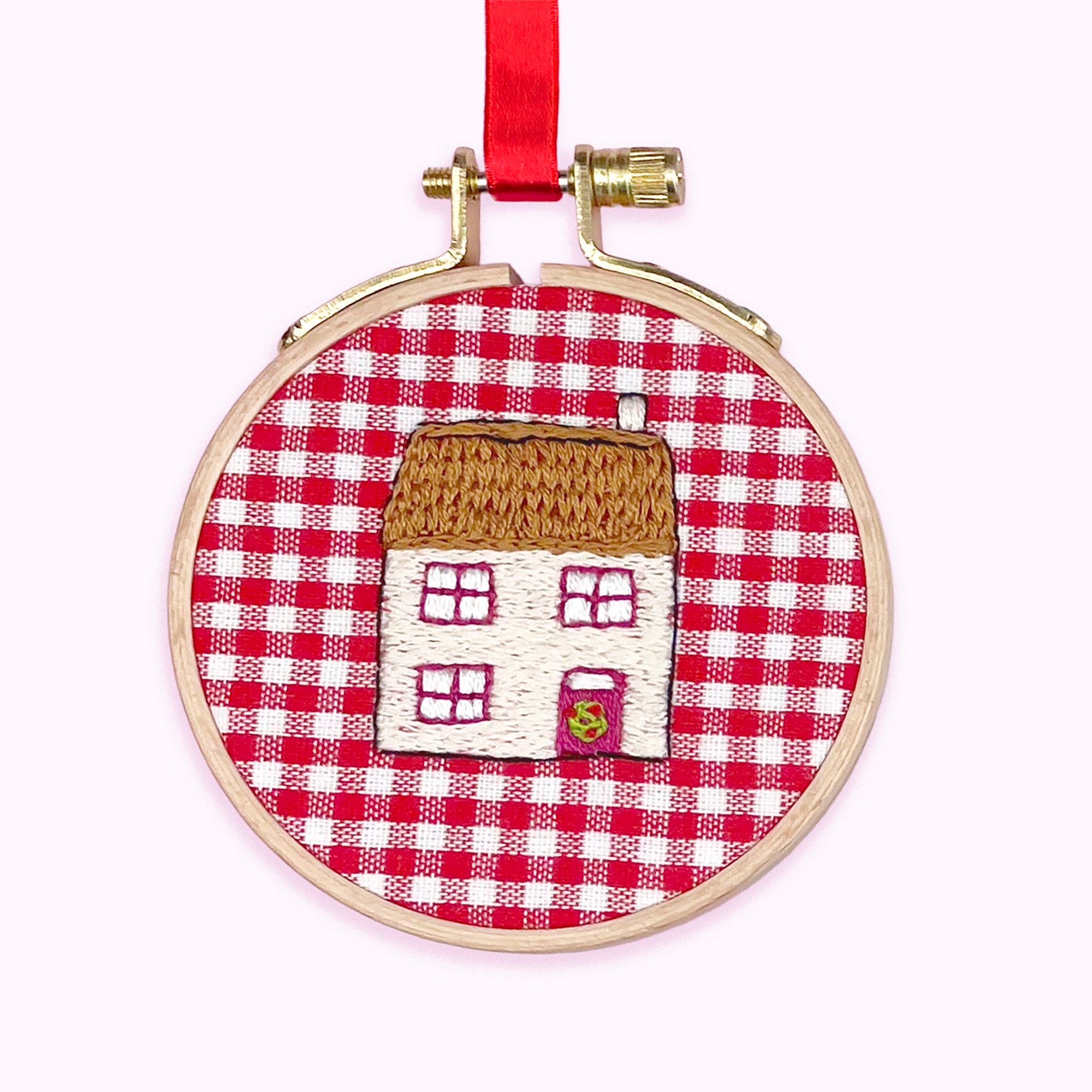 Embroidery hoop with a house design on red and white checkered fabric, hanging on a white background.