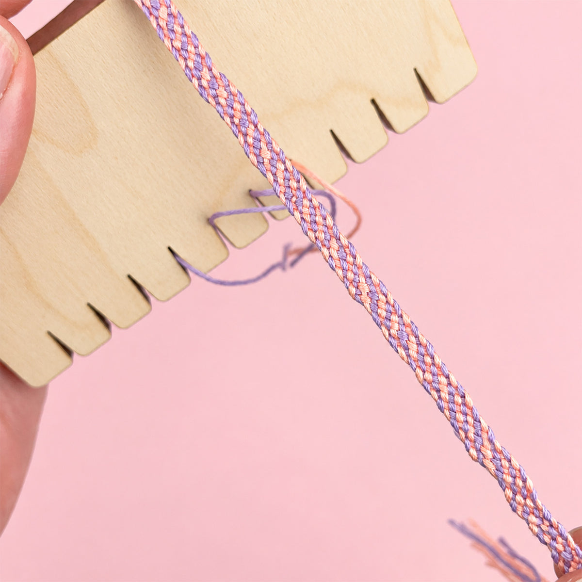 A close-up of a hand holding a wooden braiding tool and a braided friendship bracelet with purple, pink, and orange threads against a solid pink background.
