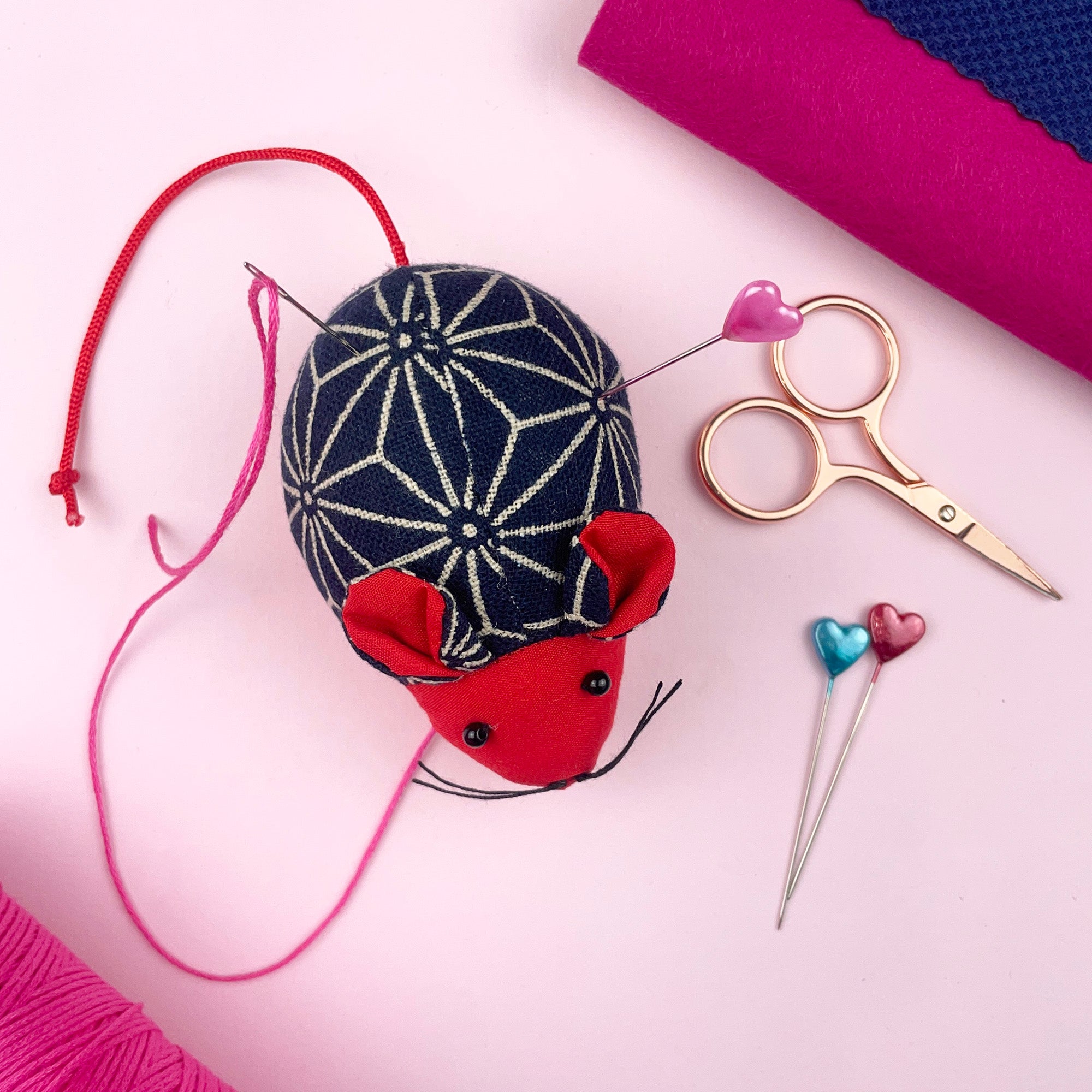 Decorative mouse pin cushion with sewing tools on a pink background. Mouse pin cushion in red and blue fabric with whiskers and heart-topped pins, a fun sewing accessory.