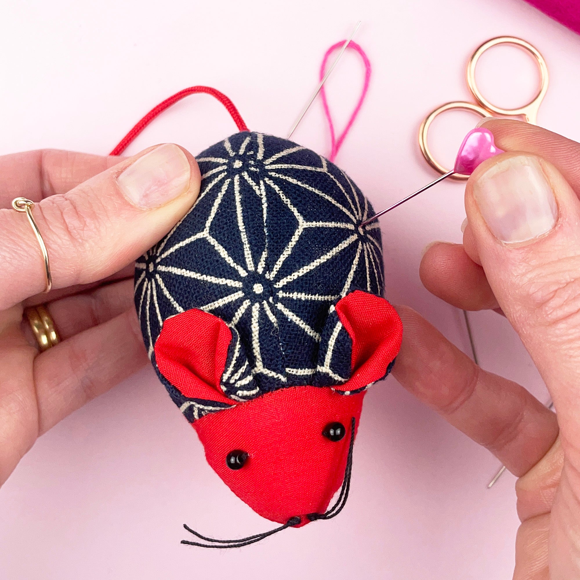 Small mouse-shaped sewing pin cushion in patterned navy fabric with red details, used for holding needles.
