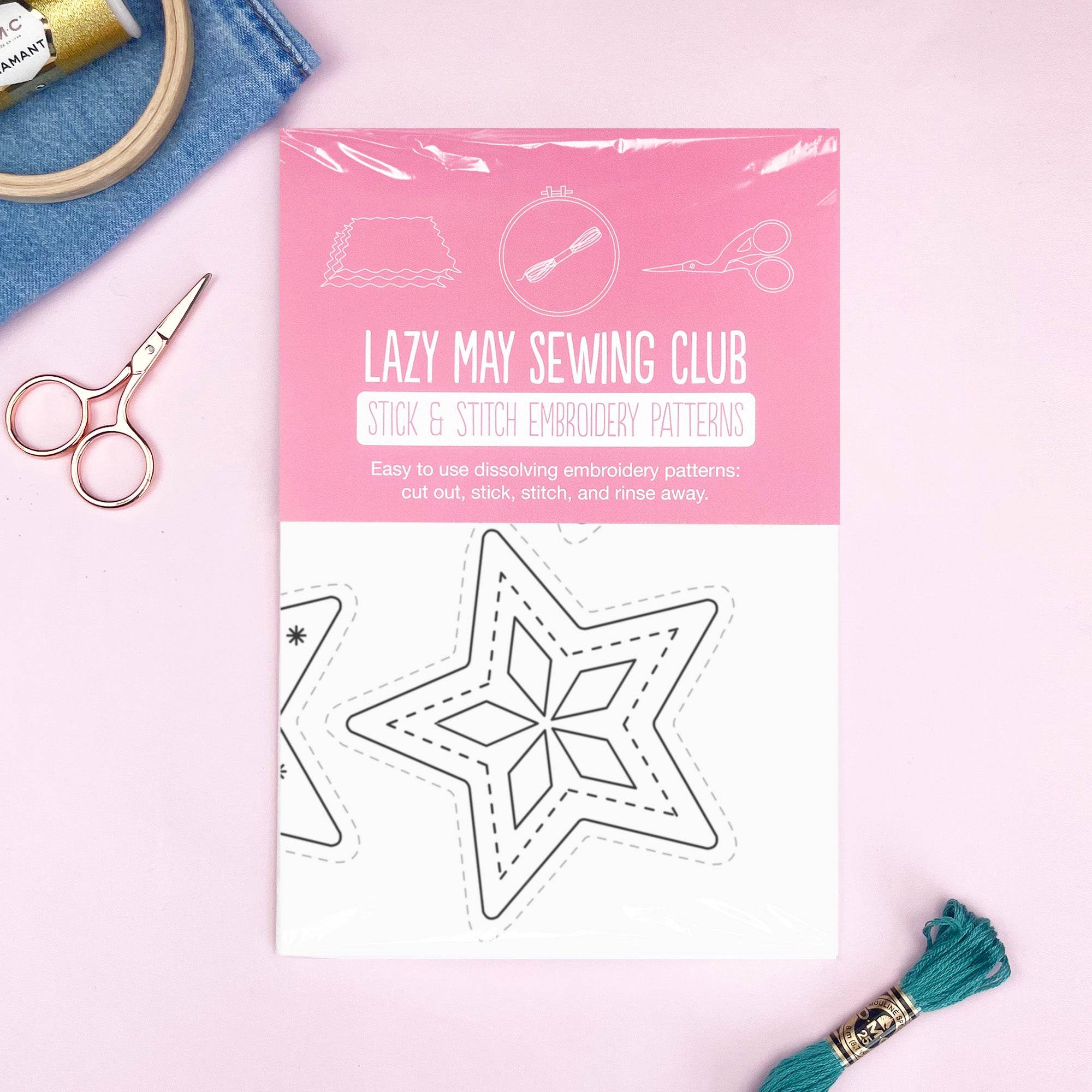 A packaged embroidery pattern booklet from Lazy May Sewing Club lies on a pink surface, surrounded by small crafting props including scissors, a spool of thread, denim fabric, and a tassel. The package is pink and white with line drawings and the text 'Lazy May Sewing Club Stick & Stitch Embroidery Patterns.