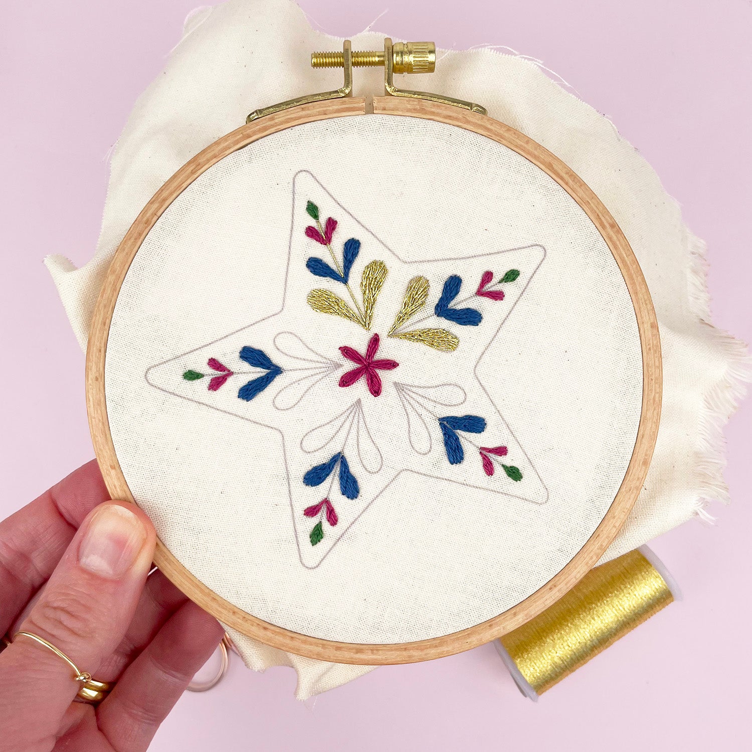 A hand holds a wooden embroidery hoop containing white fabric with a star-shaped floral embroidery design in blue, green, and pink thread. A spool of gold thread rests on a light pink background.