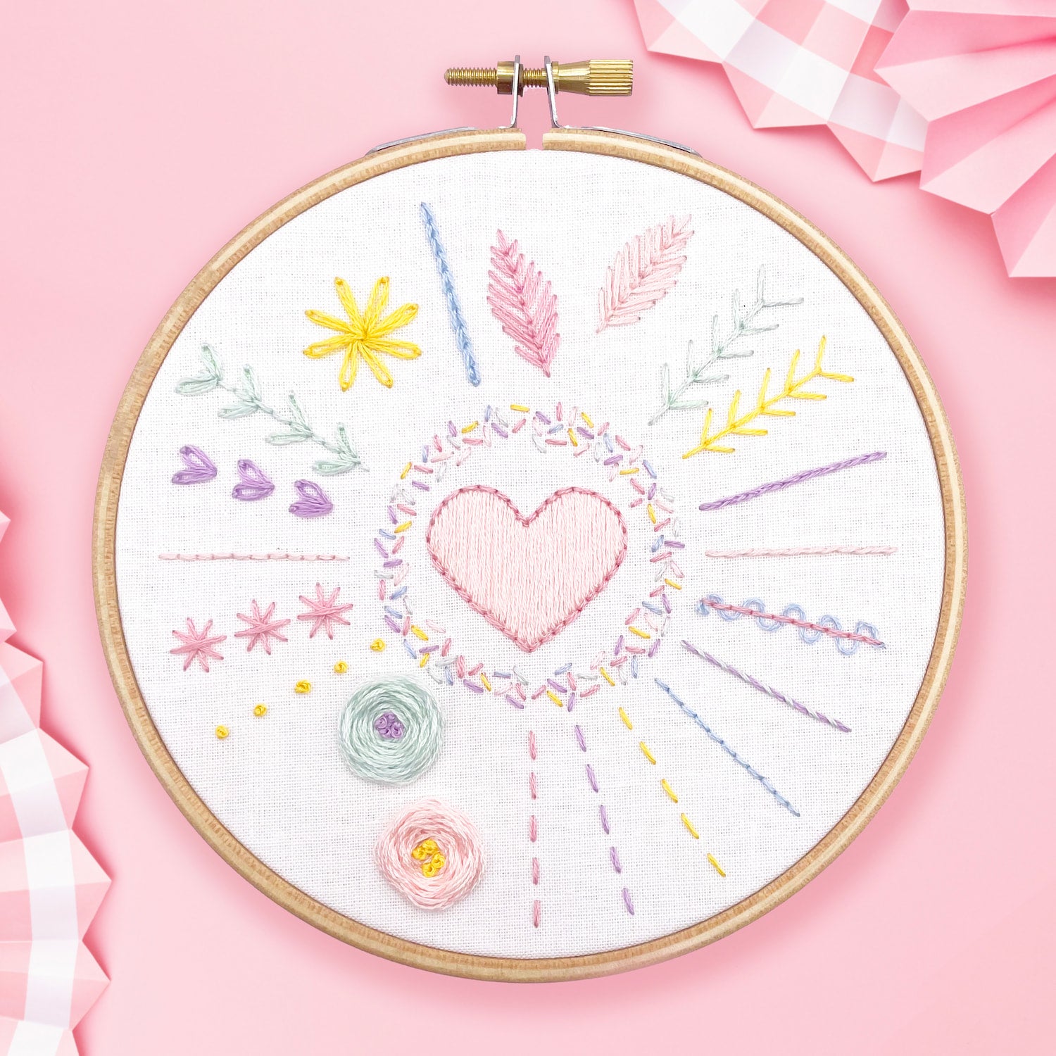 Embroidery hoop with colorful stitches and heart design from Embroidery Kit: 5" Beginners Stitch Sampler on pink background