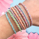 A series of pastel-colored woven friendship bracelets displayed on a wrist, with a wooden weaving loom and colourful cords visible in the background. Kumihimo style bracelets made from a friendship bracelet making kit