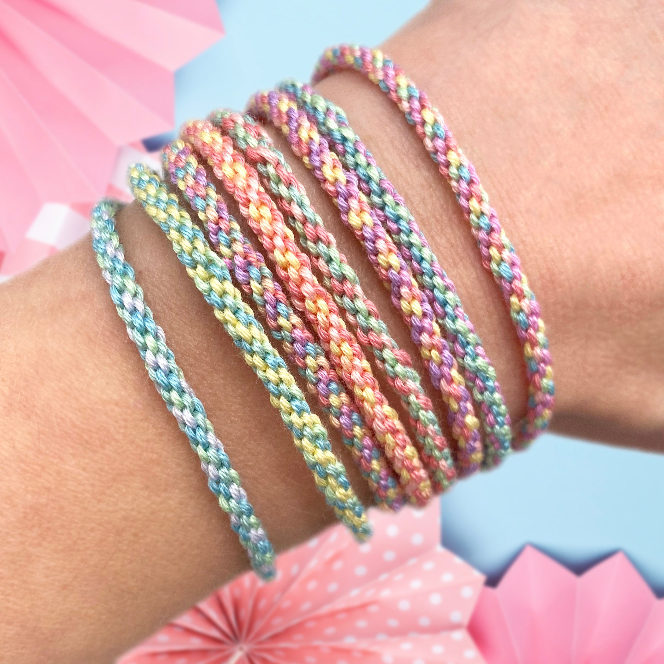 A series of pastel-colored woven friendship bracelets displayed on a wrist, with a wooden weaving loom and colourful cords visible in the background. Kumihimo style bracelets made from a friendship bracelet making kit