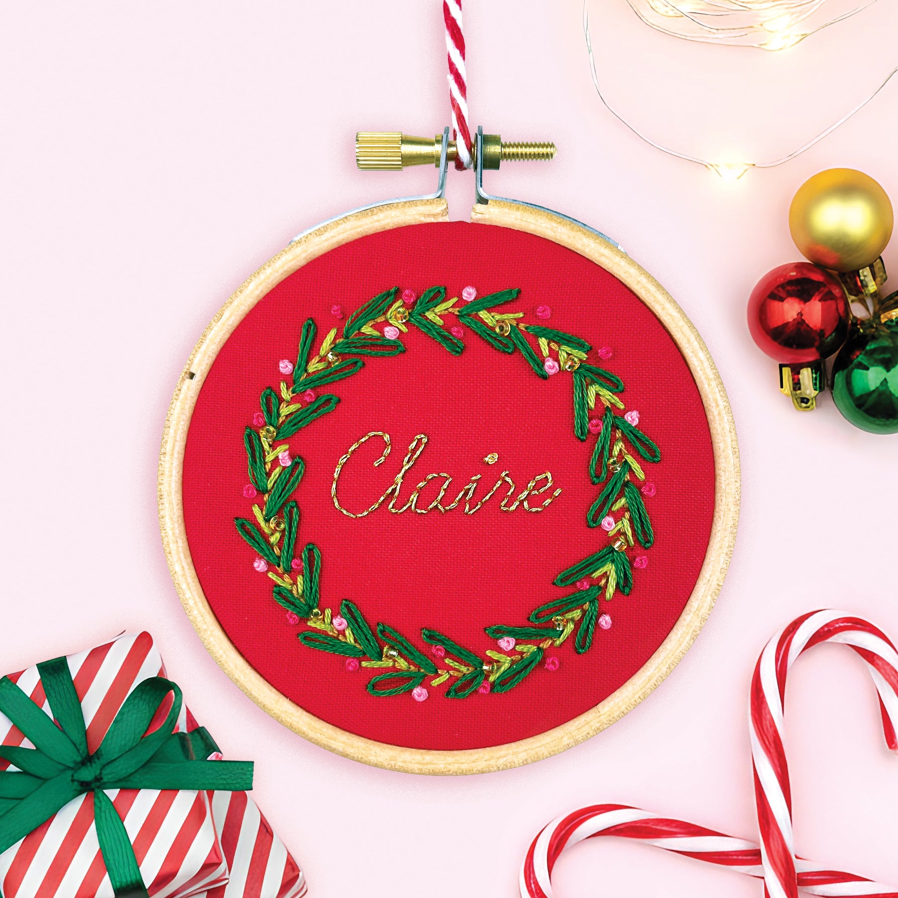 Embroidery hoop with 'Claire' design on red fabric, surrounded by Christmas decorations.
