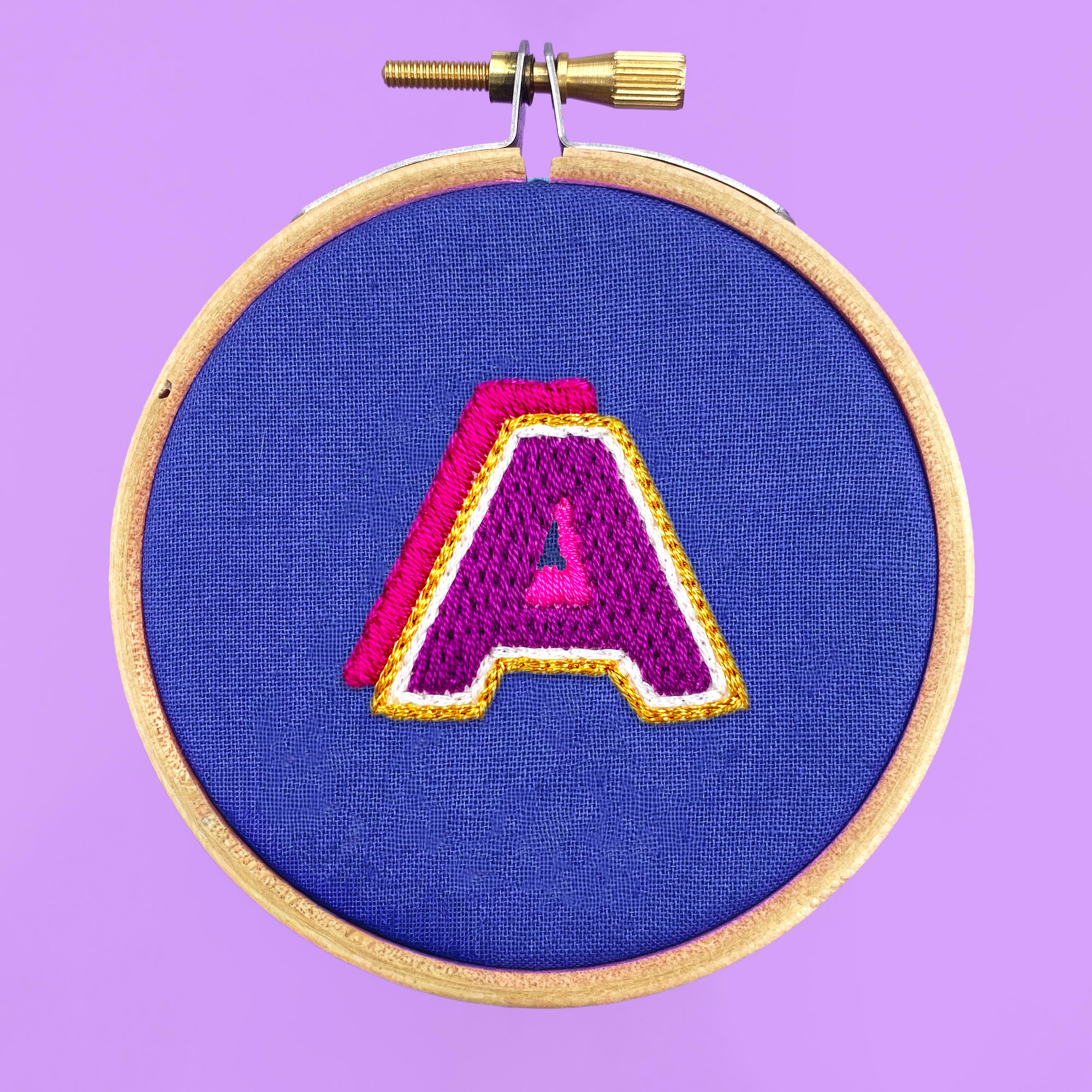 Iron-On Embroidery Patterns: 26 Bold Alphabet Letter Designs featuring colorful stitched letter A on blue fabric in hoop.