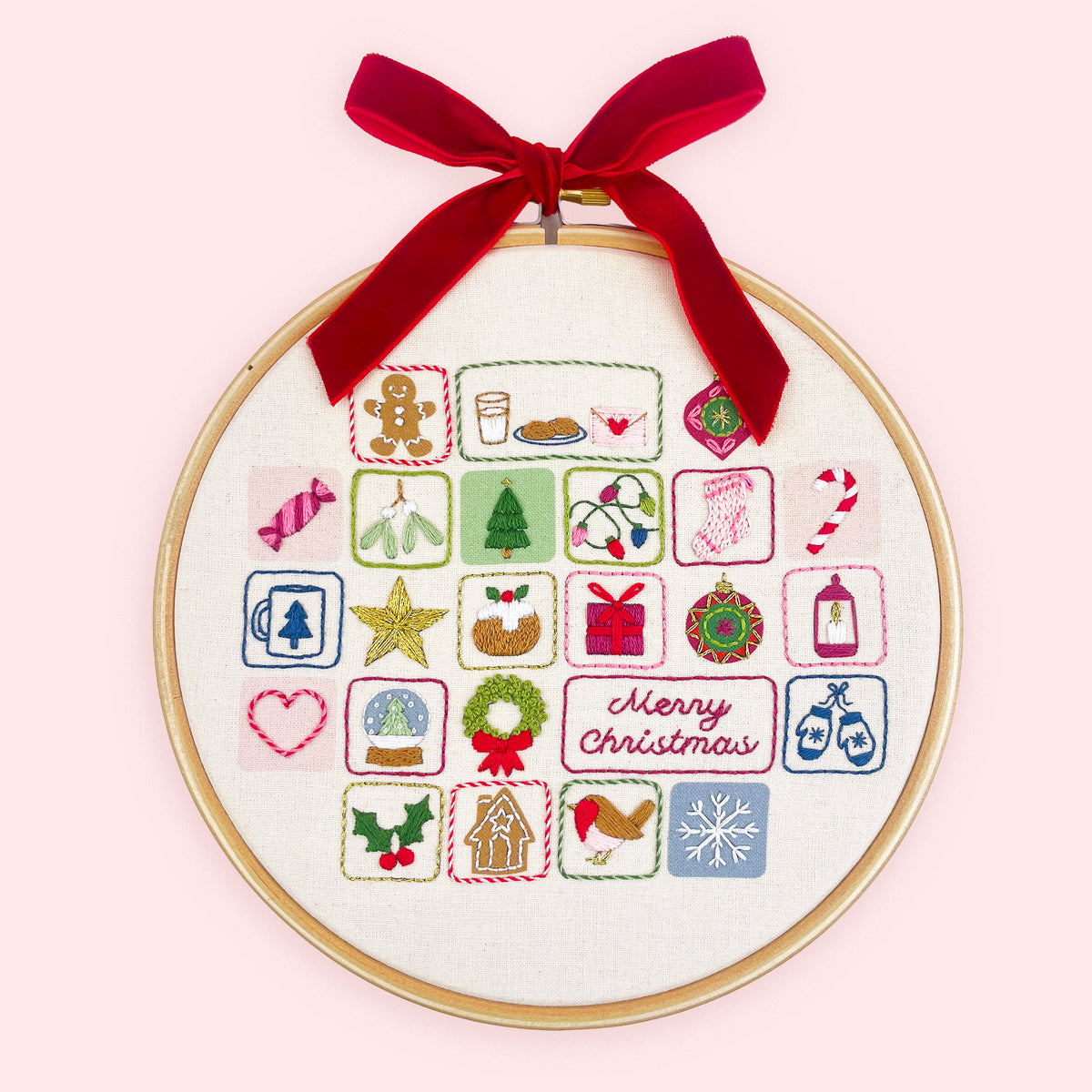 Embroidery advent calendar. A circular embroidery hoop with a wooden frame holds a white fabric panel featuring colorful Christmas-themed stitched icons and a red 'Merry Christmas' script. A red velvet ribbon is tied in a bow at the top for hanging.