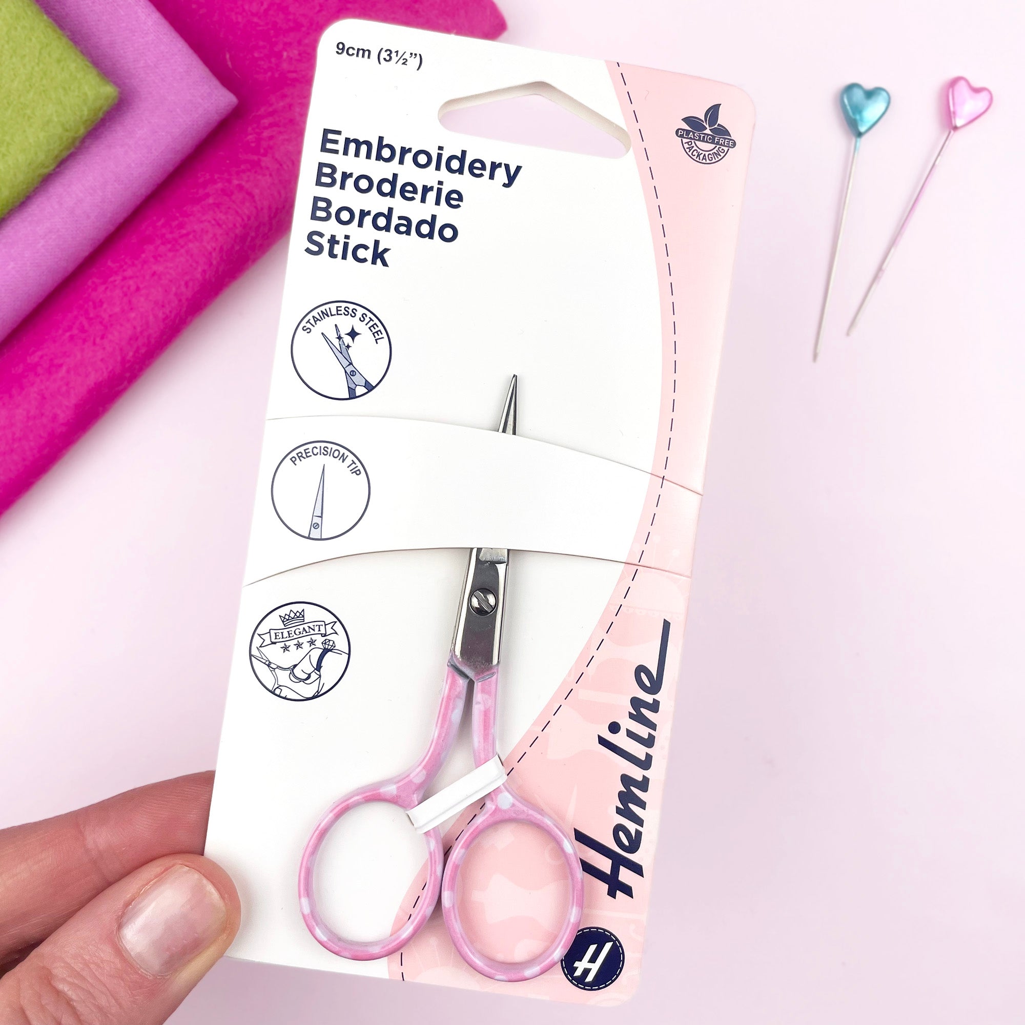Packaged embroidery scissors with pink handles held by a hand, against a light background.