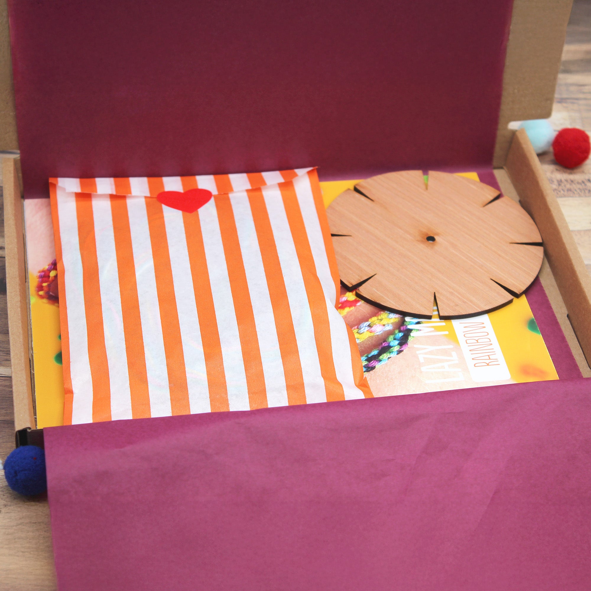 An open Lazy May rainbow bracelet kit box lined with orange-striped tissue paper, containing a wooden kumihimo loom, instruction card and colourful threads.